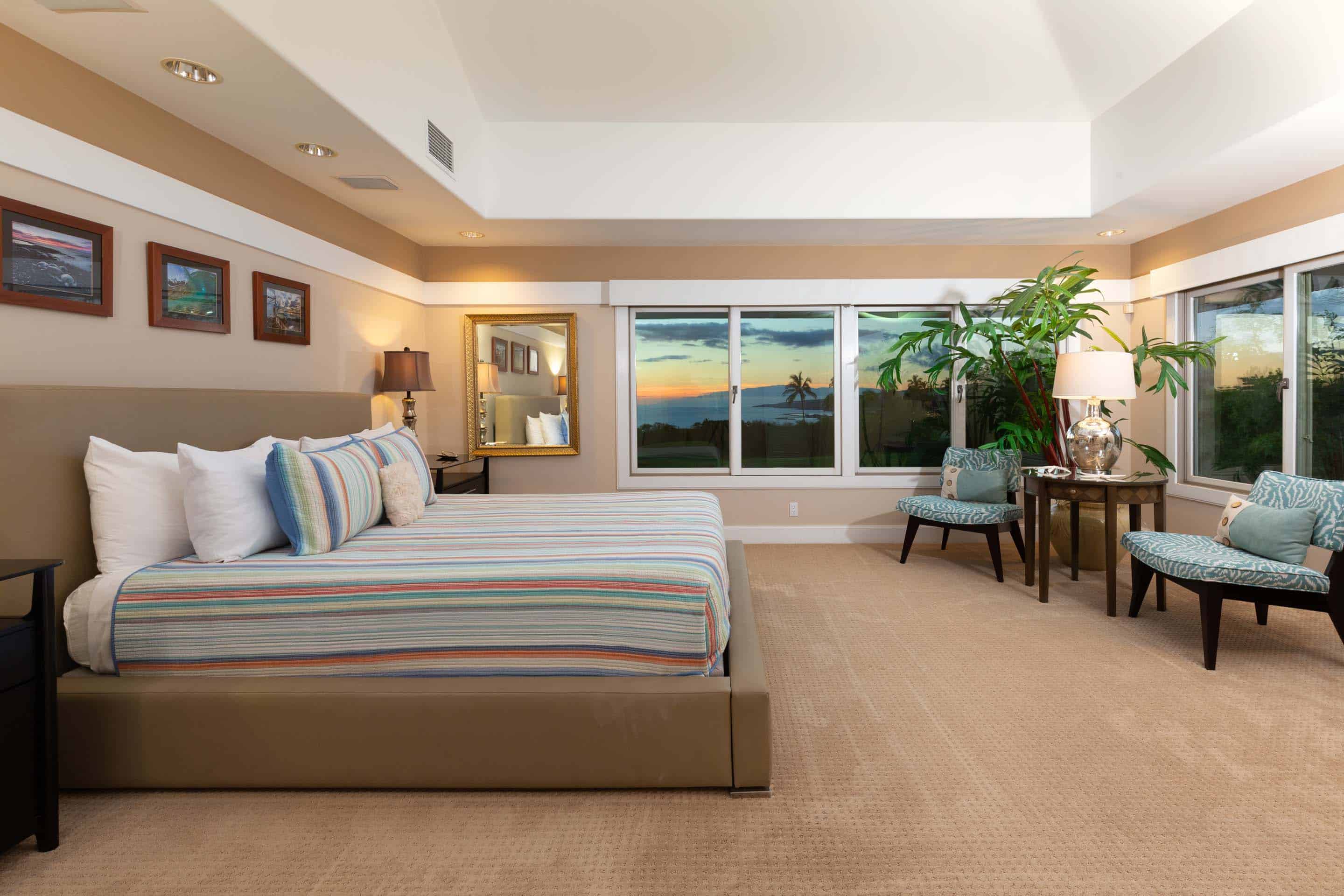 This is a spacious primary bedroom featuring a king-size bed with a striped comforter and several pillows. Large windows offer a view of the sunset over the ocean, and the room is decorated with a plant, two chairs, and a small table. The overall impression is luxurious and relaxing.