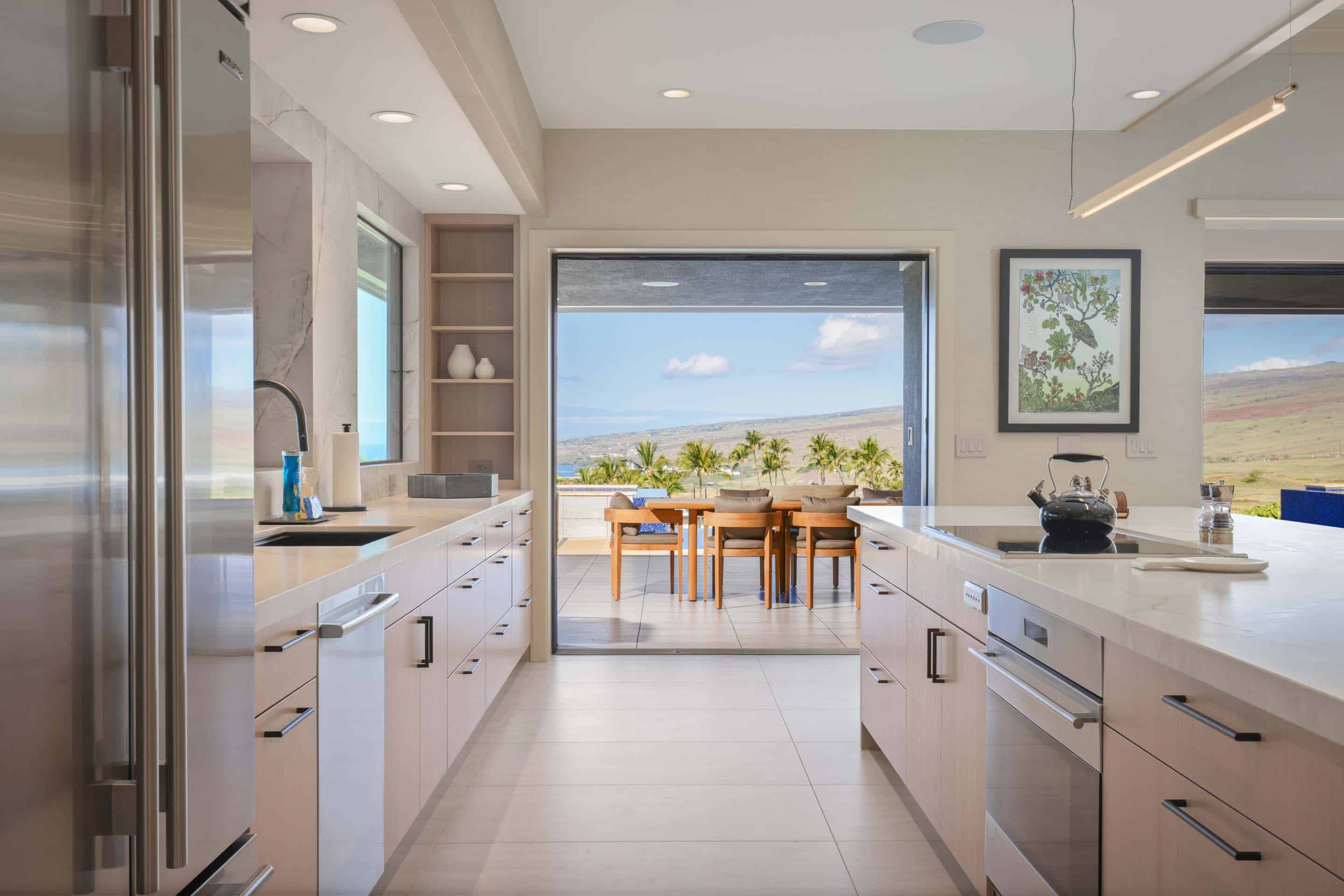 This is a bright and modern kitchen with sleek, light-colored cabinetry and stainless steel appliances. The kitchen opens to an outdoor patio area with a dining table, offering a seamless indoor-outdoor living experience. The view from the patio includes palm trees, the ocean, and a distant hillside, enhancing the property's appeal.