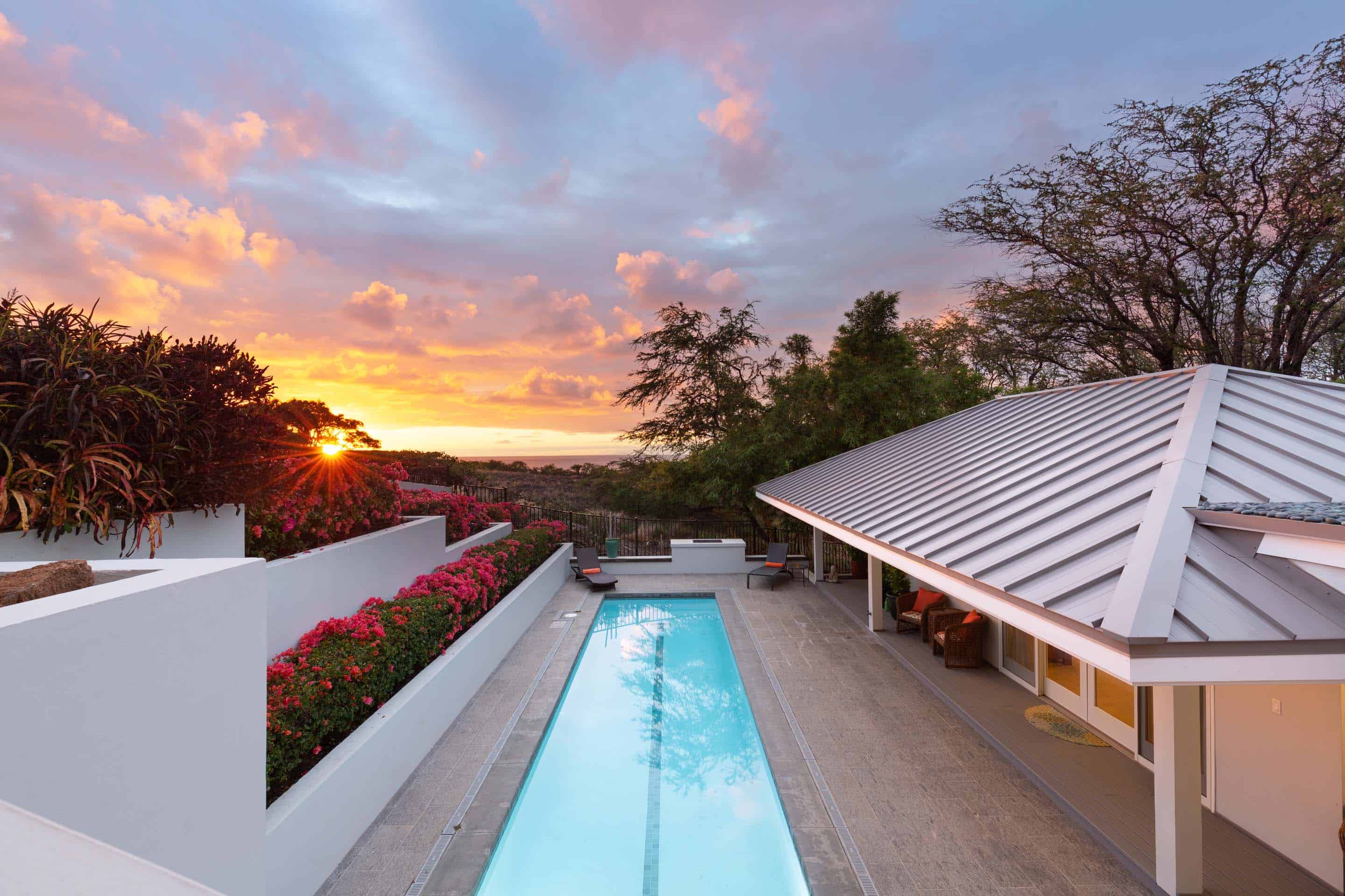 This image showcases a luxurious outdoor pool area at sunset. The rectangular pool is surrounded by a tiled patio and bordered by white walls with vibrant pink bougainvillea. A modern house with a light-colored metal roof is visible on one side, and the setting sun creates a warm and inviting atmosphere, perfect for relaxation and entertainment.