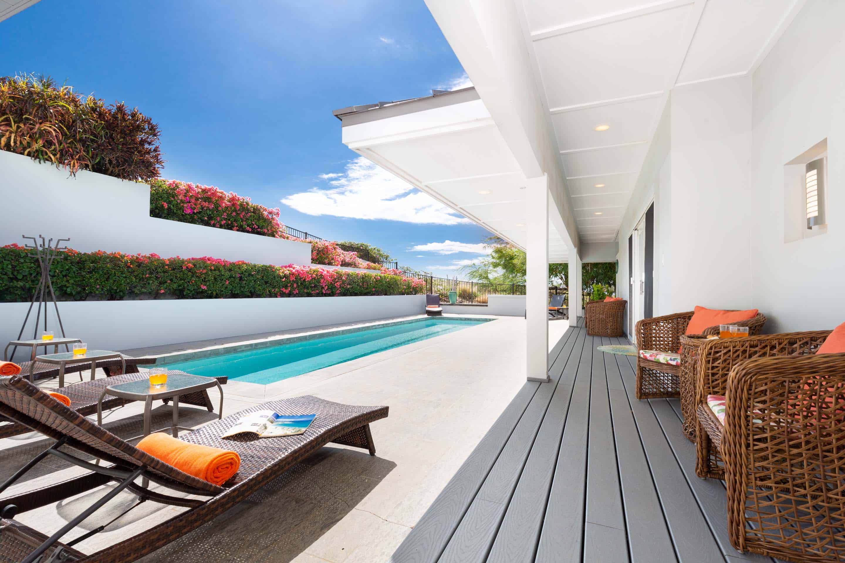 This image showcases a luxurious outdoor pool area with a sleek, modern design. The pool is surrounded by a spacious patio with lounge chairs and side tables, perfect for relaxation. A covered deck area with wicker furniture provides a shaded retreat, enhancing the overall appeal of this upscale outdoor living space.