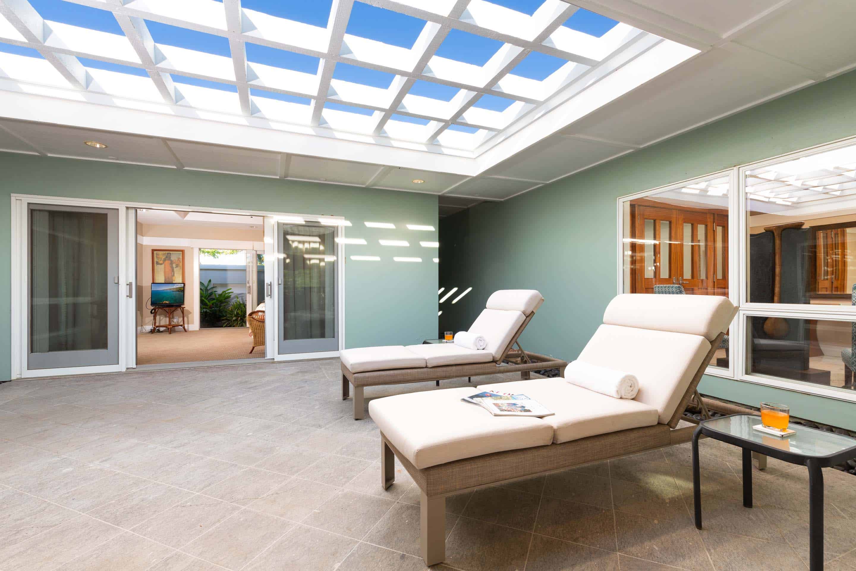 This sunroom features a tiled floor, light green walls, and a skylight with a grid pattern. Two lounge chairs with white cushions are positioned in the center of the room, with a small glass-topped table nearby. Sliding glass doors lead to another room, creating a seamless transition between indoor and outdoor spaces.