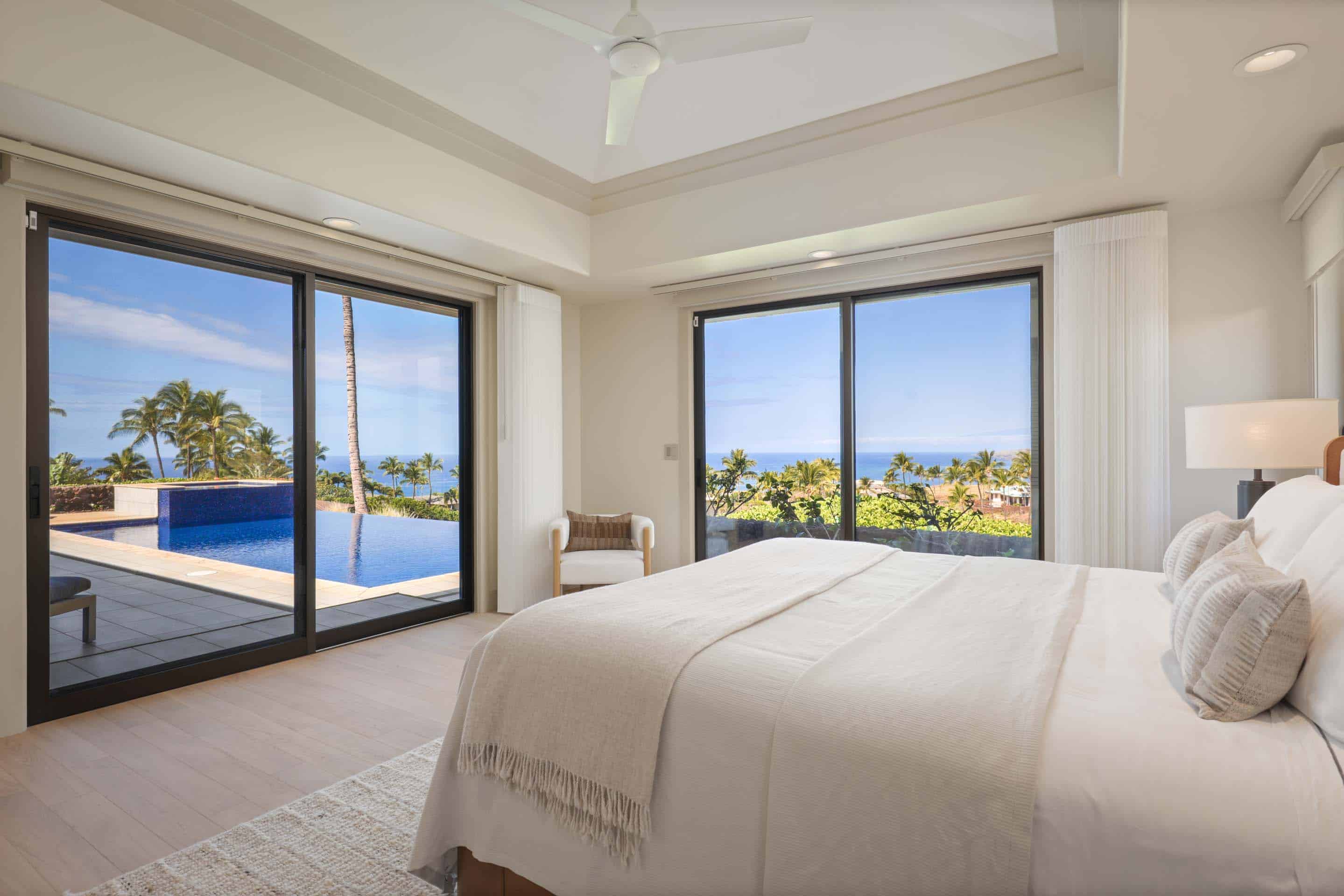 This is a luxurious primary bedroom featuring a large bed with white linens and a throw blanket. The room has floor-to-ceiling windows that offer stunning views of the ocean and a private pool. The overall aesthetic is bright, airy, and modern, creating a serene and inviting space.