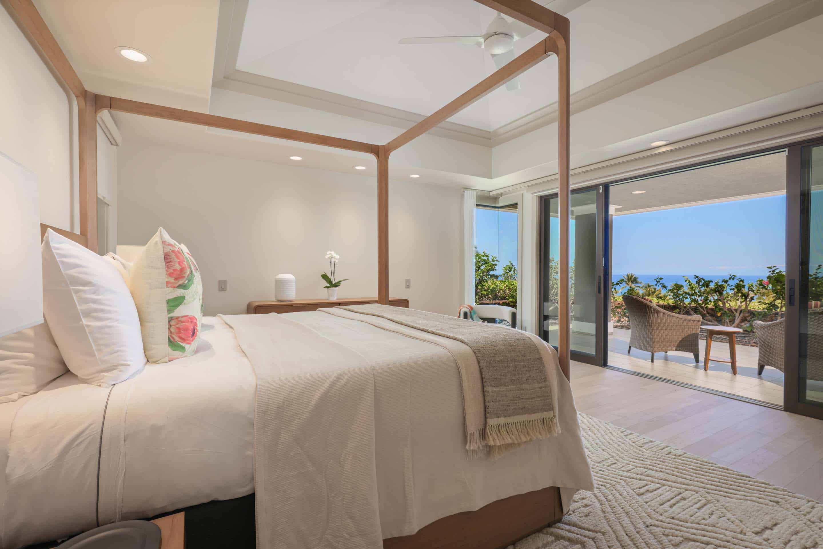 This is a bright and airy primary bedroom featuring a four-poster bed with white linens and decorative pillows. Sliding glass doors open to a patio with ocean views, creating a seamless indoor-outdoor living experience. The room is decorated in a minimalist style with light wood floors and neutral tones, enhancing the sense of tranquility and luxury.
