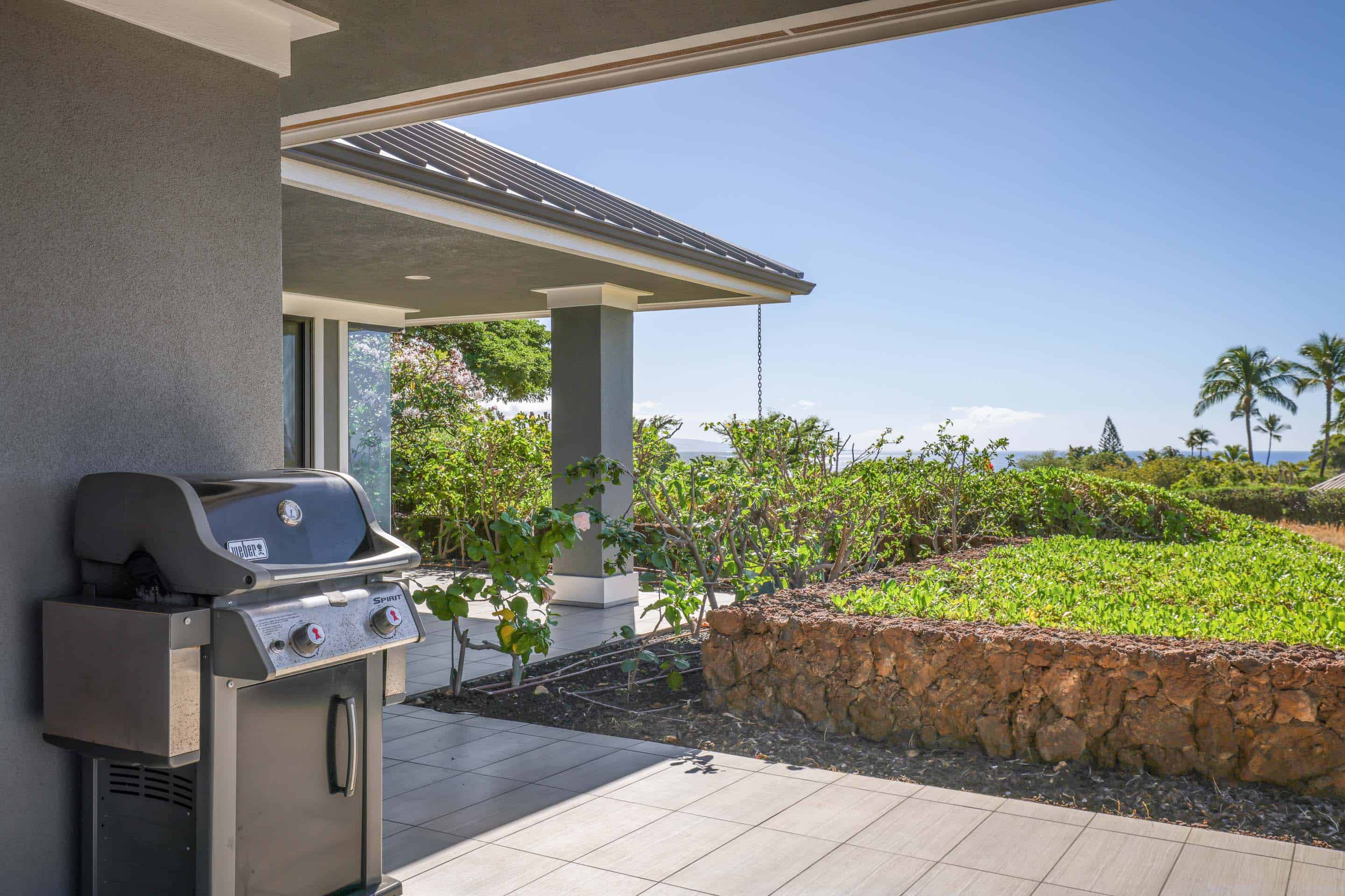 This image showcases a covered patio area with a built-in grill, offering an outdoor living space with a view. The patio features tiled flooring and a low stone wall bordering a garden area, with glimpses of the ocean and palm trees in the distance. The overall impression is one of relaxed luxury and outdoor enjoyment.