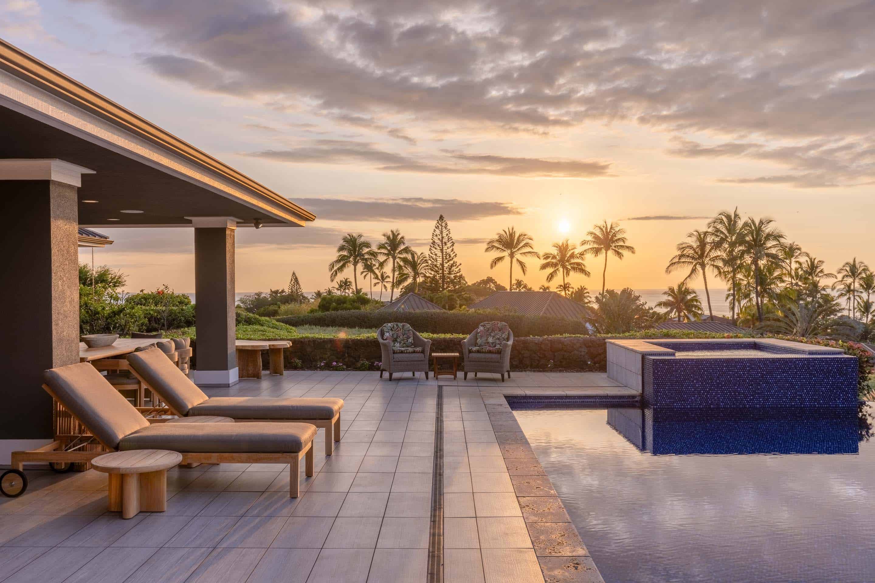This image showcases a luxurious outdoor pool and spa area at sunset. The pool features a dark blue tiled spa section, and is surrounded by a tiled patio with lounge chairs and seating. Palm trees and lush landscaping create a tropical backdrop, enhancing the property's appeal as a relaxing and upscale retreat.