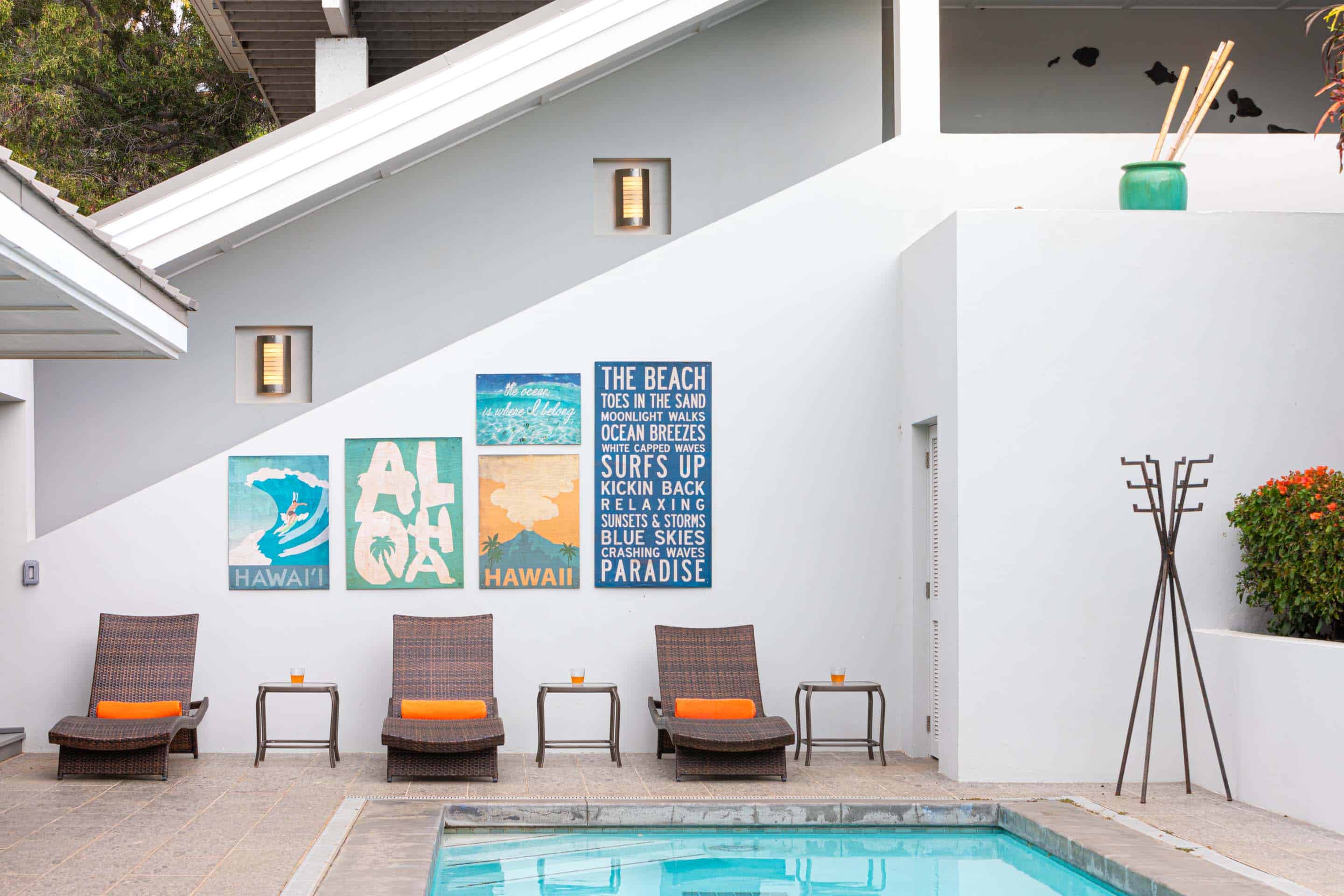 This image showcases a serene pool area with a modern architectural backdrop. Rattan lounge chairs with orange cushions are arranged alongside small tables, creating a relaxing poolside setting. The white walls are adorned with beach-themed artwork, adding a touch of coastal charm to the outdoor space.