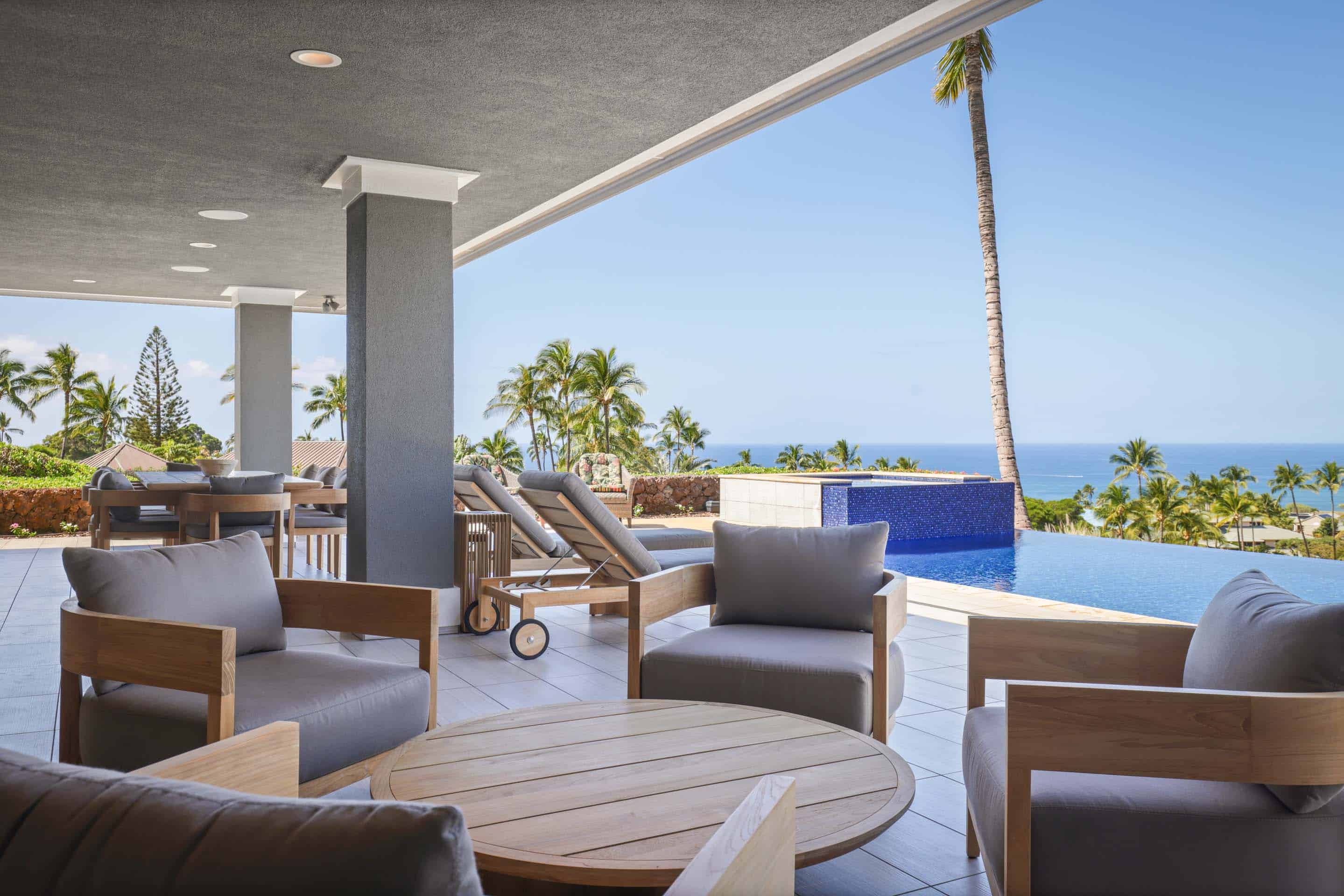 This image showcases a luxurious outdoor patio area with comfortable seating arrangements, including armchairs and lounge chairs, arranged around a wooden coffee table. The patio overlooks an infinity pool with a stunning ocean view framed by palm trees, creating a serene and upscale ambiance. The architectural design features clean lines and modern finishes, enhancing the property's appeal.