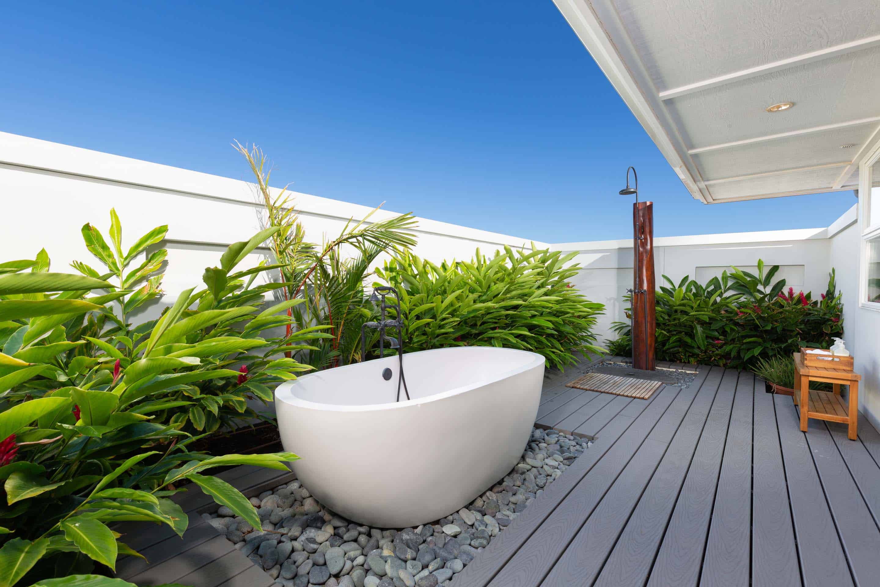 This image showcases a luxurious outdoor bathroom space, featuring a freestanding white bathtub set on a bed of smooth stones, surrounded by lush tropical greenery. A wooden deck extends to an outdoor shower, creating a spa-like atmosphere under a clear blue sky. The perspective is a medium shot, highlighting the integration of nature and modern design.
