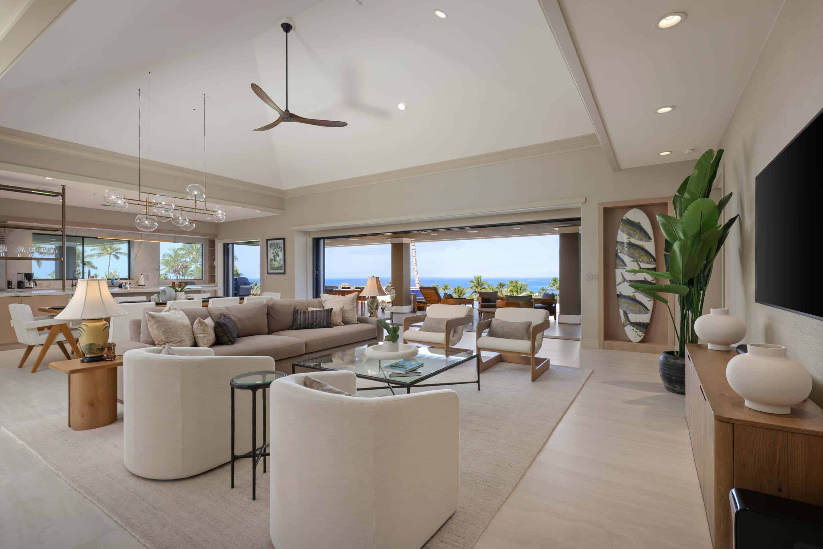This is a bright and airy living room with a modern coastal design. The space features a large sectional sofa, white armchairs, and a glass coffee table, all arranged on a light-colored rug. Large sliding glass doors open to an ocean view, enhancing the indoor-outdoor living experience.