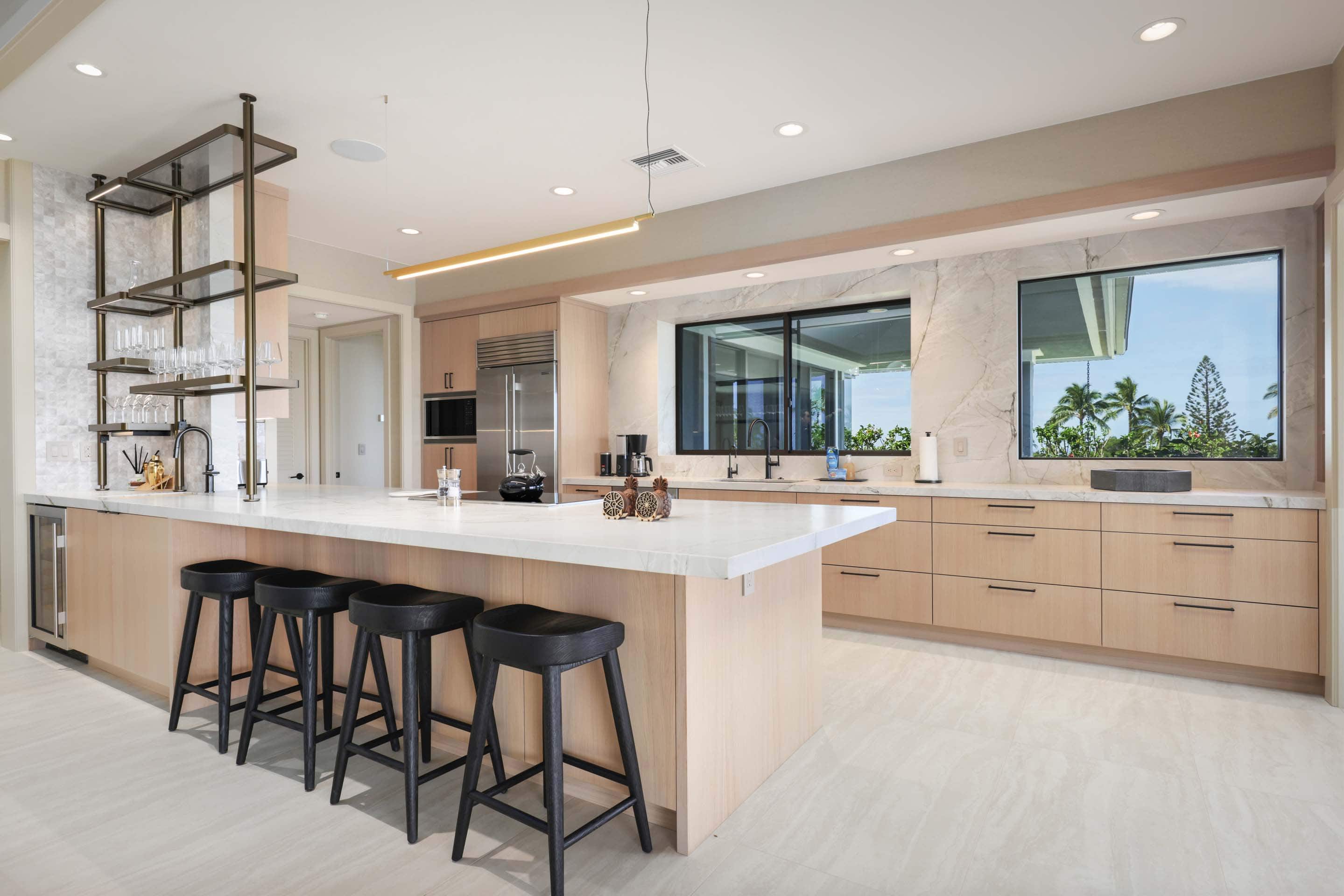 This is a bright and modern kitchen featuring light wood cabinetry, a large white countertop island with black stools, and stainless steel appliances. The kitchen is well-lit with recessed lighting and a linear pendant light above the island. Large windows provide natural light and views of the outdoors, enhancing the open and airy feel of the space.