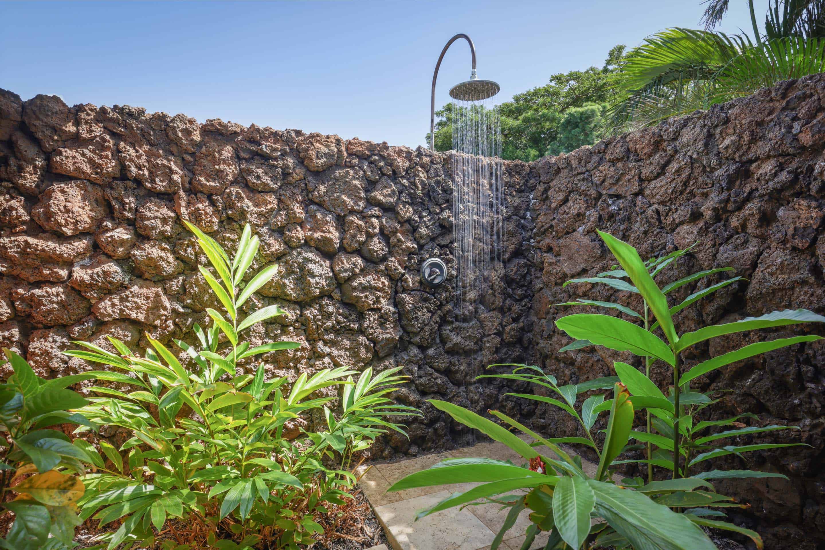 This image showcases an outdoor shower area enclosed by lava rock walls, creating a private and natural setting. Lush green plants surround the shower, adding to the tropical ambiance. The shower features a rainfall showerhead, suggesting a luxurious and refreshing experience.
