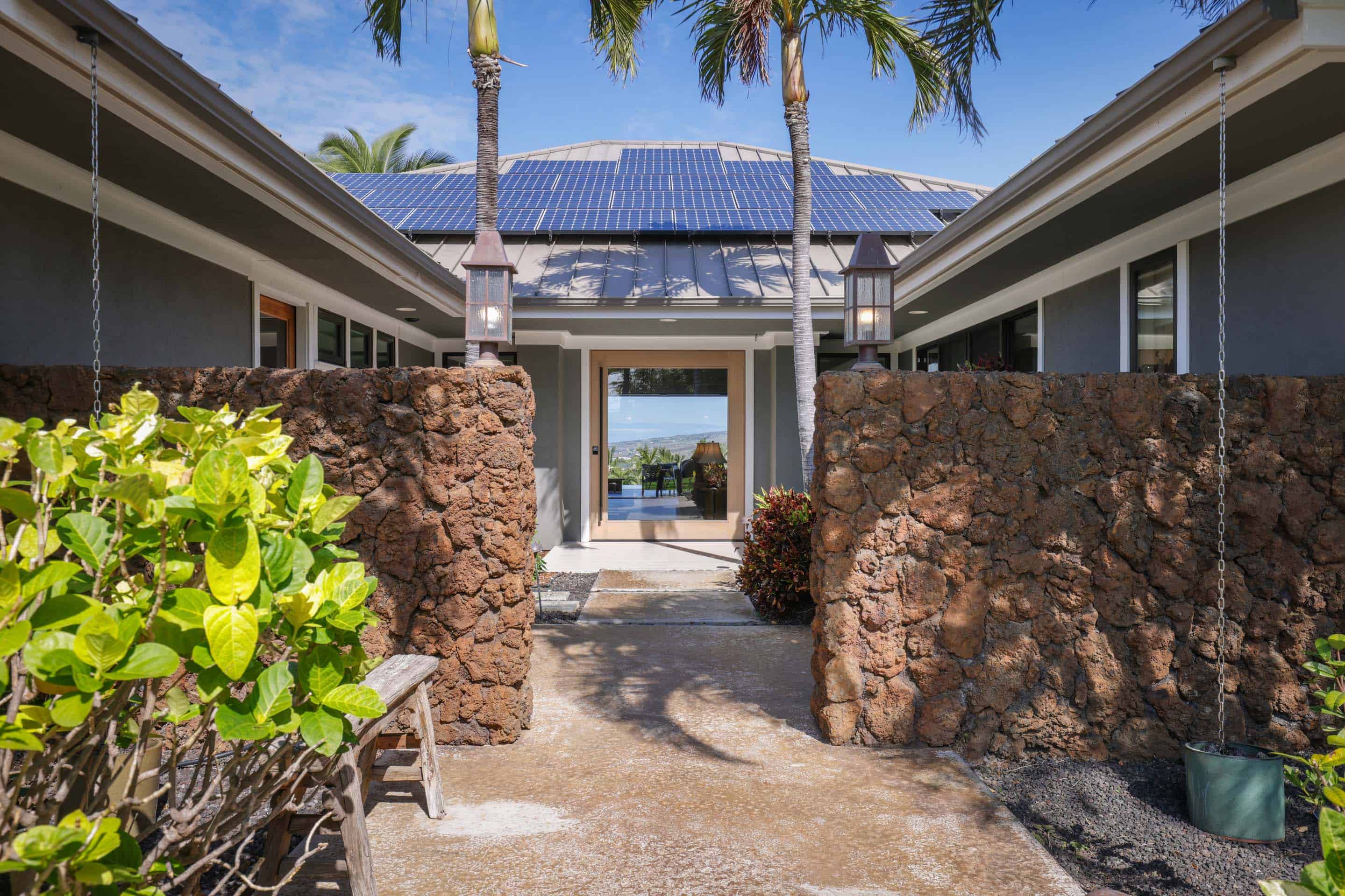 This is an inviting entryway to a modern home, featuring lava rock walls flanking a pathway leading to a glass-paneled front door. Solar panels are visible on the roof above the entrance, and palm trees add a tropical touch. The perspective is from the pathway, looking towards the house.