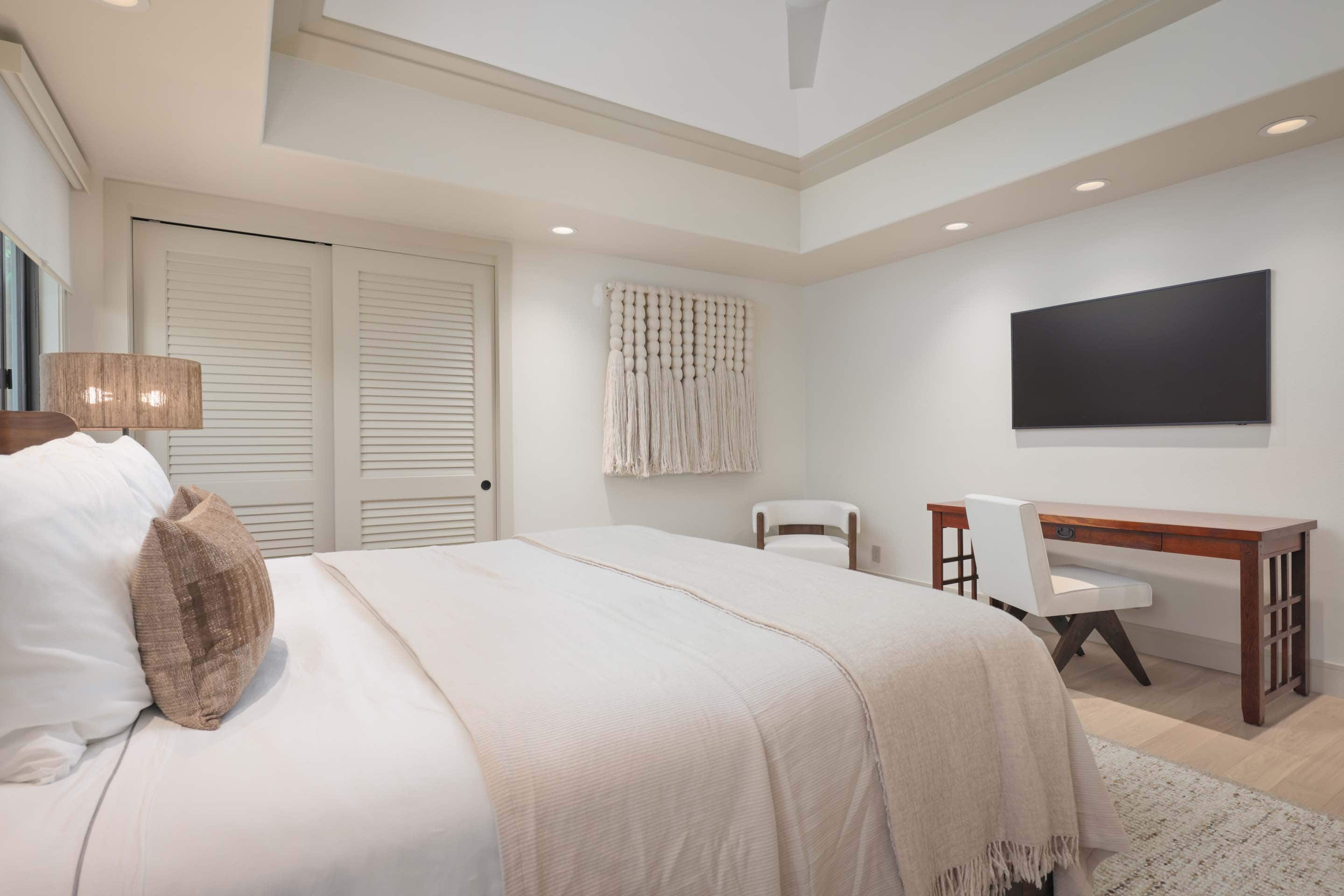 This is a bright and airy primary bedroom featuring a king-size bed with white linens and a decorative throw. A wooden desk with a white chair sits beneath a wall-mounted television, and a macrame wall hanging adds a touch of texture. The room has a clean, modern aesthetic with recessed lighting and light wood floors.