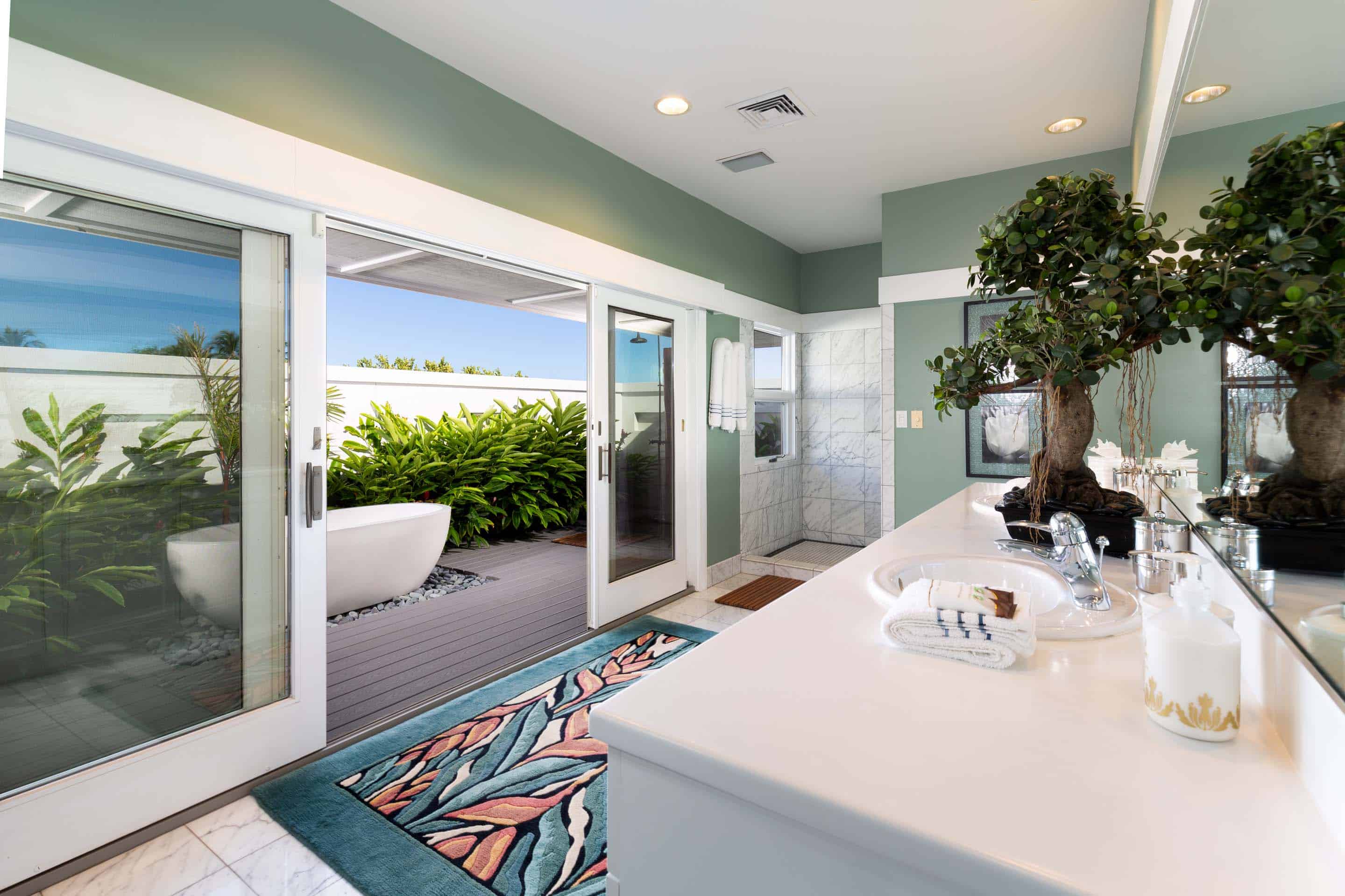 This is a bright and airy primary bathroom featuring a double vanity with white countertops, modern fixtures, and a large mirror. Sliding glass doors open to a private outdoor patio with a soaking tub surrounded by lush greenery, creating a spa-like retreat. The color palette is soothing, with light green walls and white trim, enhancing the sense of relaxation.