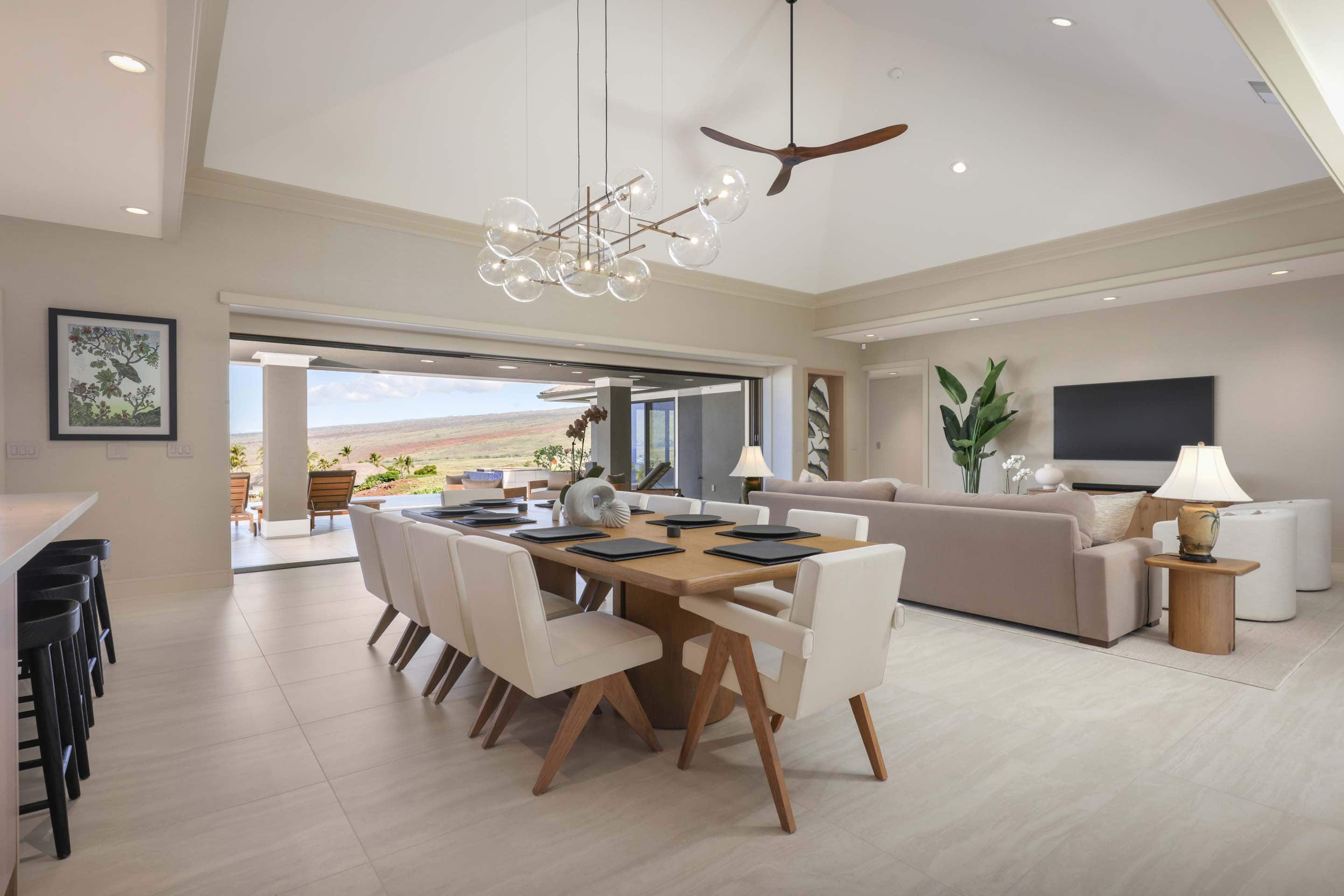 This interior shot showcases a bright and airy dining room that seamlessly blends into the living space. The dining area features a large wooden table with seating for eight, complemented by a modern bubble chandelier. Sliding glass doors open to an outdoor view, enhancing the indoor-outdoor flow, while the living area includes a comfortable sofa and a television, creating a cohesive and inviting atmosphere.