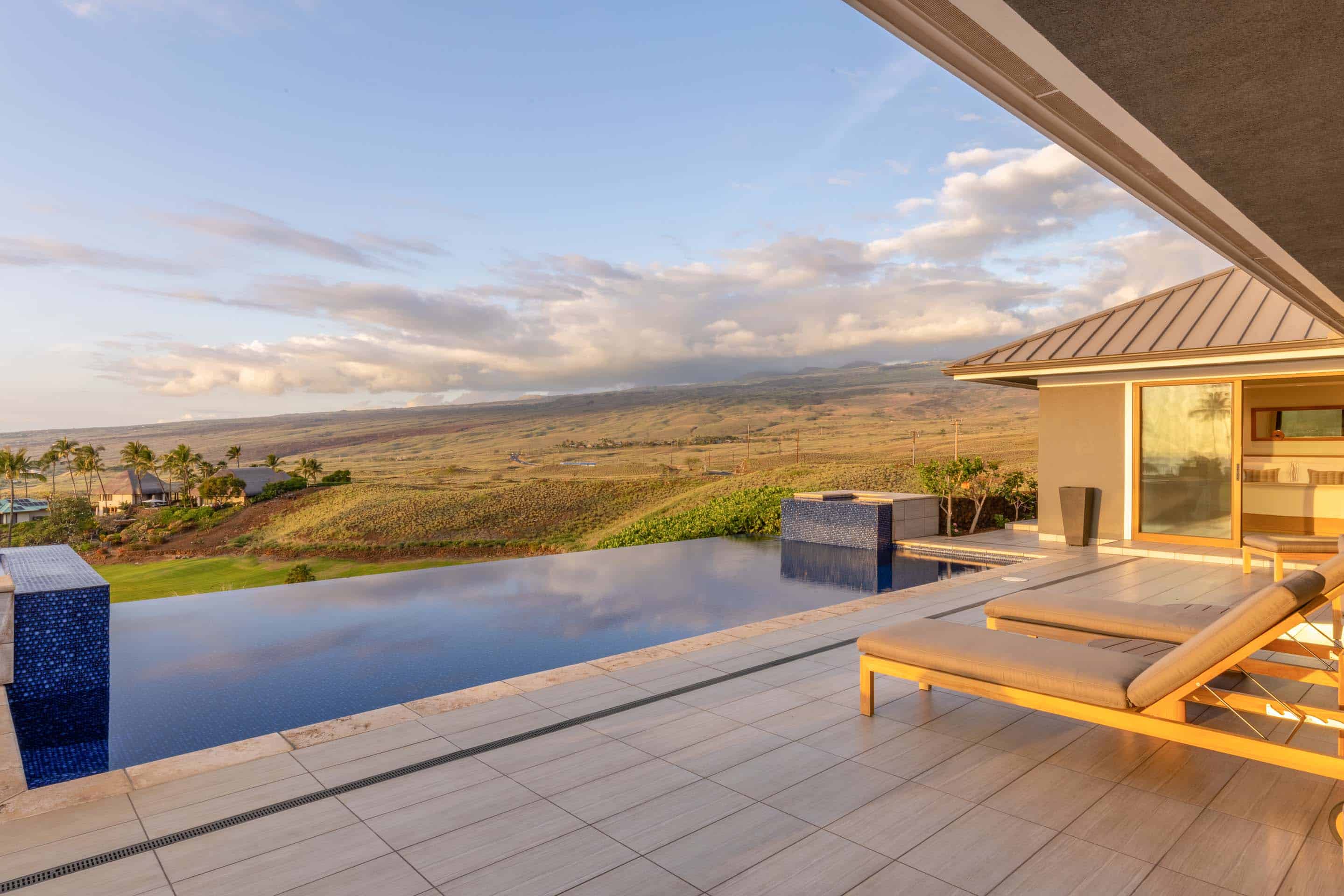This image showcases a luxurious infinity pool overlooking a vast, scenic landscape. The pool's dark blue water reflects the sky, creating a seamless transition between the pool and the horizon. Two lounge chairs are positioned on the tiled patio, inviting relaxation and enjoyment of the stunning view from this high-end property.
