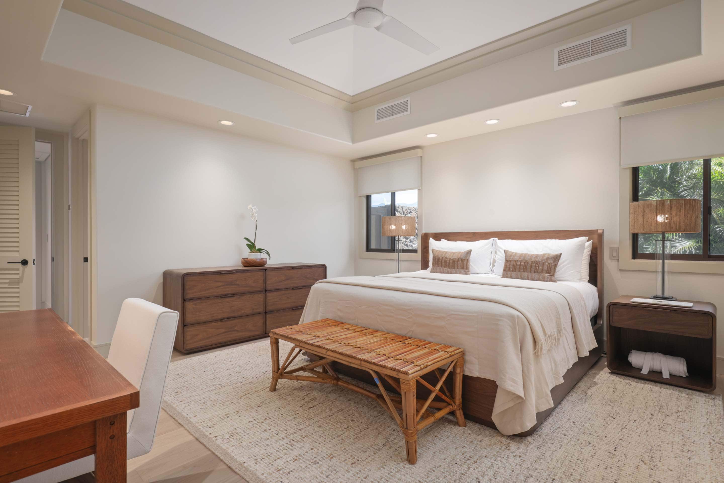This is a well-lit primary bedroom featuring a wooden bed frame with neutral bedding, complemented by a woven bench at the foot of the bed. A wooden dresser and nightstand add to the room's warm tones, while a large area rug softens the space. The room is decorated with minimalist decor, including an orchid and lamps, creating a serene and inviting atmosphere.