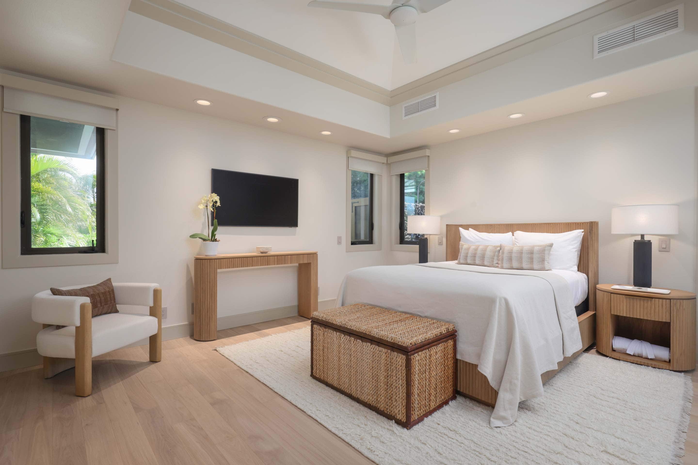 This is a bright and airy primary bedroom featuring a king-size bed with a woven headboard, a woven storage bench at the foot of the bed, and matching nightstands. A modern console table sits beneath a wall-mounted television, and a comfortable armchair is placed near the window. The room is decorated in neutral tones with light wood flooring and a plush area rug, creating a serene and inviting atmosphere.