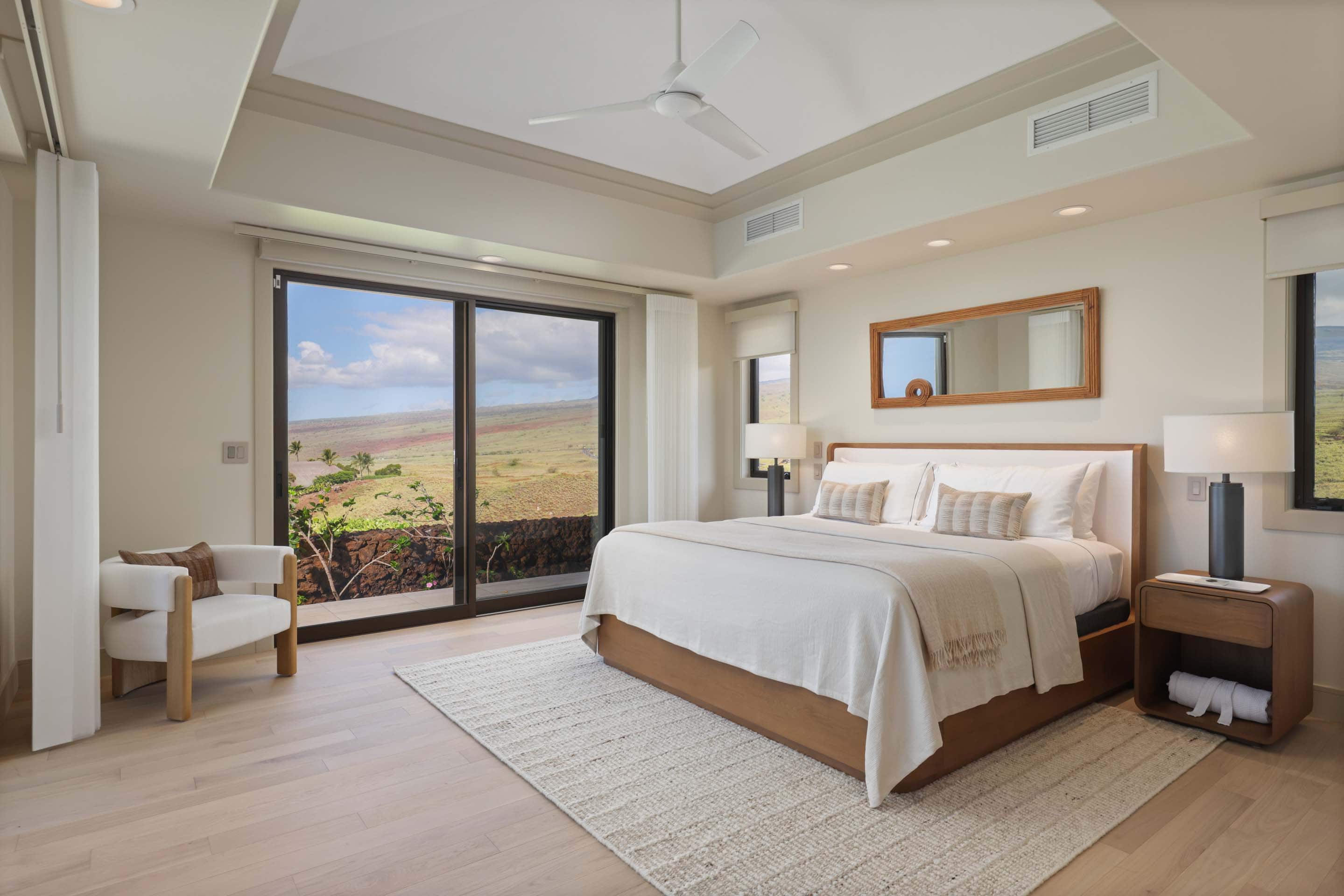 This is a bright and airy primary bedroom featuring a king-size bed with white linens and decorative pillows, a wooden nightstand with a lamp, and a comfortable armchair. Large sliding glass doors offer a stunning view of the landscape, enhancing the room's serene and luxurious atmosphere. The room is decorated in neutral tones with wood accents, creating a relaxing and inviting space.