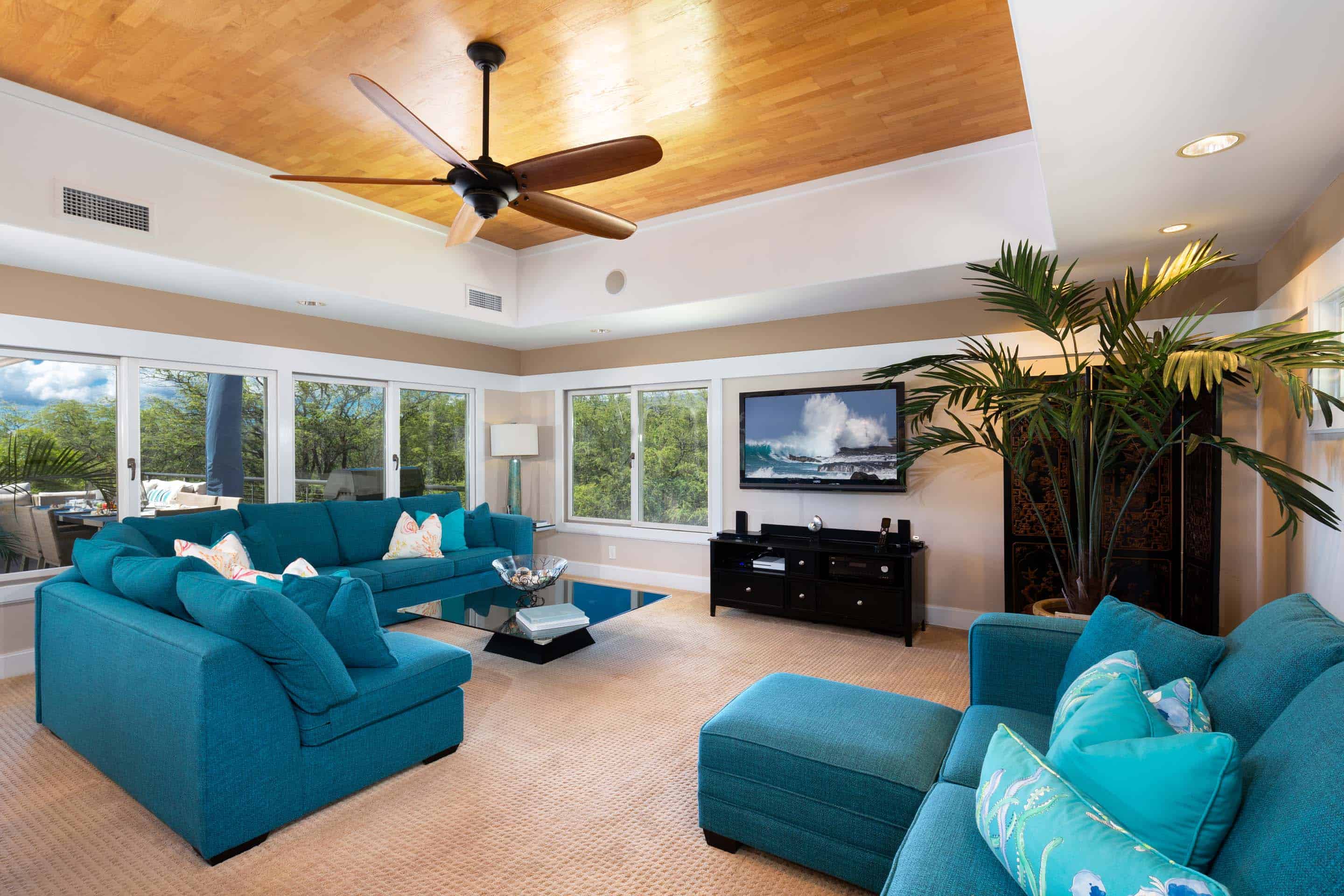 This is a bright and inviting living room featuring a large, teal sectional sofa with decorative pillows, a glass coffee table, and a dark wood entertainment center with a flat-screen TV. Large windows offer ample natural light and views of the outdoors, while a wooden ceiling and a ceiling fan add character to the space. A large potted plant adds a touch of nature to the room.