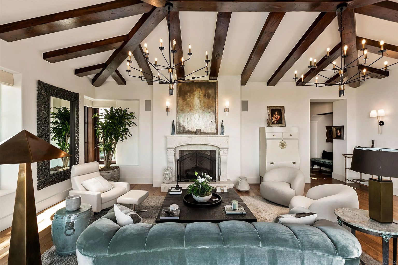 This sophisticated living room features exposed dark wood ceiling beams and a central stone fireplace with a tall, ornate surround. The space is furnished with a plush, velvet-textured sofa, two curved armchairs, and a large dark wood coffee table, all arranged on a textured area rug. Two large, multi-armed chandeliers hang from the vaulted ceiling, and a tall mirror with an intricate frame adds depth to the room, creating an elegant and inviting atmosphere.