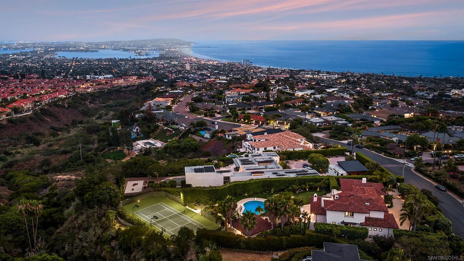 This high-angle aerial view captures a sprawling coastal estate at dusk, showcasing a large property with a private tennis court, a swimming pool, and a multi-level residence with red-tiled roofs. The perspective extends across a dense residential neighborhood toward the expansive ocean horizon under a soft, twilight sky. The scene emphasizes the property's prime location and its integration into a scenic, elevated coastal landscape.