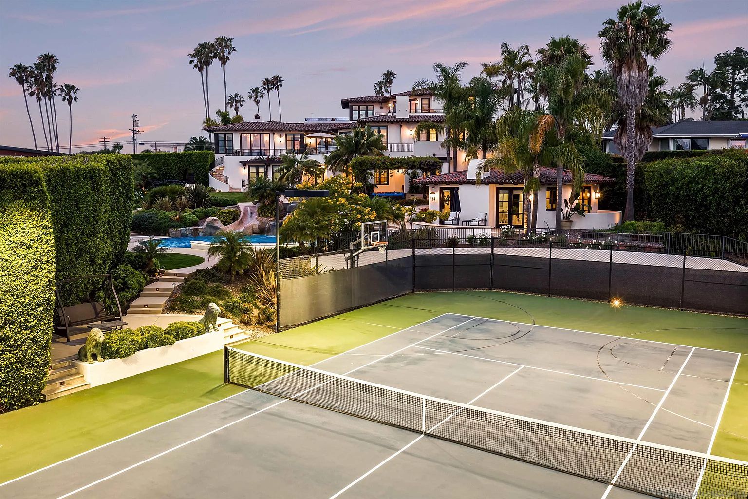 This expansive rear view captures a luxurious multi-level estate featuring a private tennis court in the foreground and a swimming pool with a slide nestled into the landscaped grounds. The Mediterranean-style residence showcases white stucco walls, dark-framed windows, and a terracotta tile roof, set against a soft sunset sky. The property is surrounded by lush greenery and mature palm trees, creating a serene and high-end resort-like atmosphere.