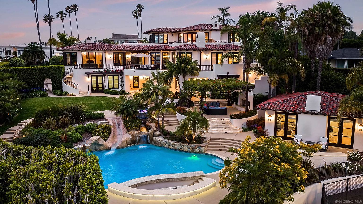 This expansive Mediterranean-style estate is captured from an elevated rear perspective, showcasing a multi-level residence with a terracotta tile roof and white stucco facade. The backyard features a resort-style pool with a rock waterfall and integrated slide, surrounded by lush landscaping, palm trees, and a stone-paved patio area. A covered outdoor living space with a pergola and a separate pool house structure complete this luxurious, private oasis.