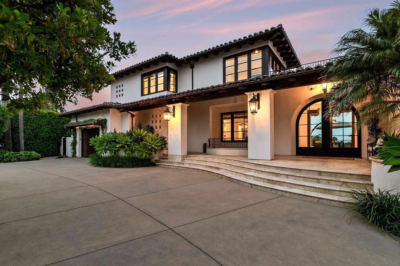 This elegant Mediterranean-style home features a crisp white stucco facade, dark-framed windows, and a classic terracotta tile roof. A wide, curved stone staircase leads up to a covered porch area, which is accented by warm wall-mounted lanterns and lush landscaping. The perspective captures the expansive concrete driveway and the inviting, symmetrical architecture during the soft light of dusk.