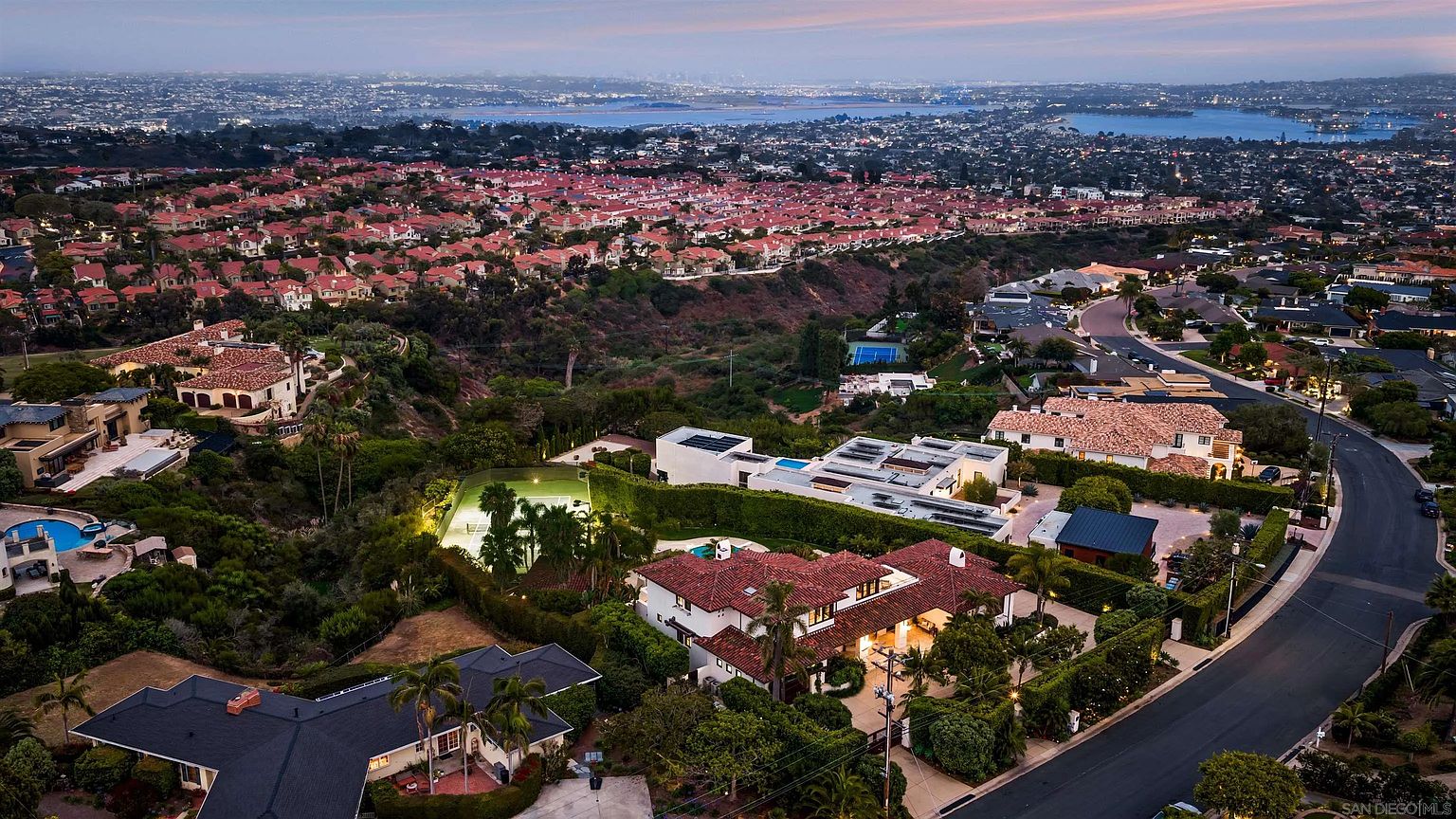This high-angle aerial view captures a sprawling residential neighborhood at dusk, featuring several large, multi-story homes with terracotta-tiled roofs. The scene highlights a prominent estate in the foreground with a private tennis court and a swimming pool, set against a backdrop of rolling hills and a distant bay. The perspective provides a comprehensive look at the property's layout, landscaping, and its integration into the surrounding suburban landscape.