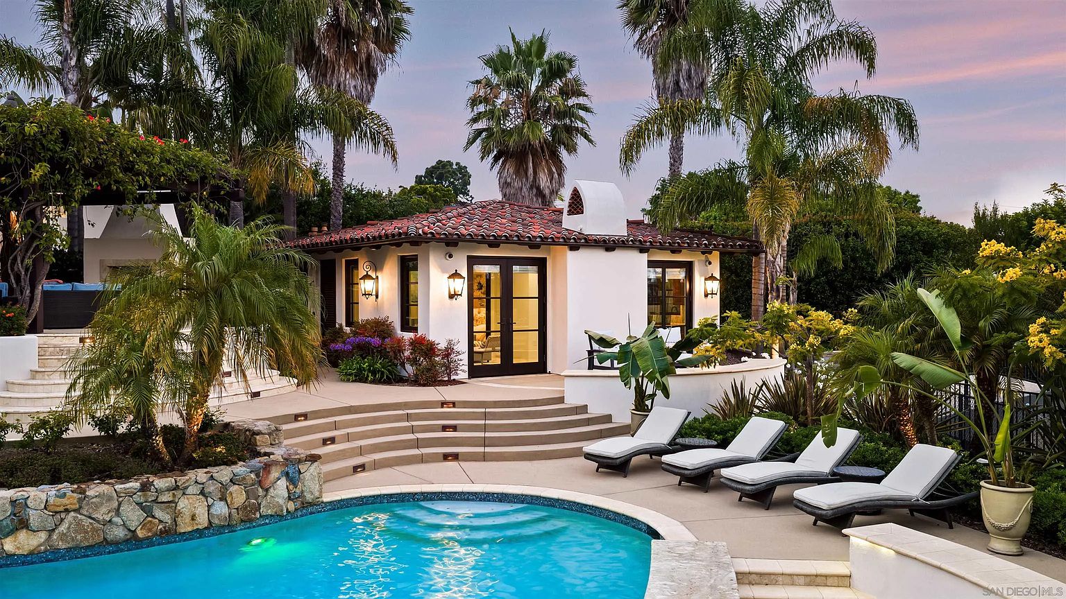 This inviting backyard scene features a curved swimming pool set against a Mediterranean-style pool house with a terracotta tile roof and white stucco walls. A tiered concrete patio area provides ample space for relaxation, furnished with four lounge chairs positioned to overlook the water. Lush tropical landscaping, including tall palm trees and vibrant greenery, frames the space, creating a serene and private resort-like atmosphere.