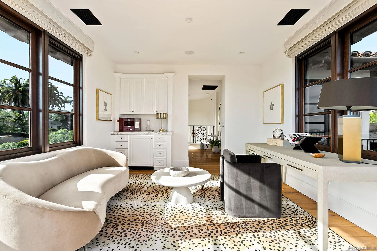 This sophisticated home office or study features a curved cream-colored sofa, a central pedestal coffee table, and a sleek desk paired with a dark upholstered chair. The room is anchored by a bold animal-print area rug and is illuminated by natural light streaming through large windows on both sides. A built-in white cabinetry unit with a countertop serves as a convenient refreshment station, while the open doorway leads to a bright hallway with a decorative metal railing.