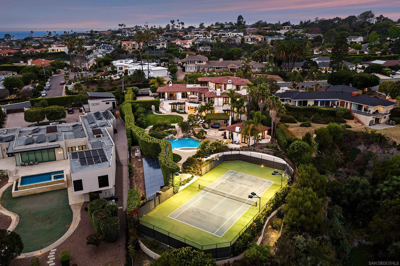 This high-angle aerial view captures a sprawling luxury estate featuring a Mediterranean-style main residence with a red-tiled roof and a private tennis court illuminated by floodlights. The property includes a landscaped backyard with a swimming pool, a water slide, and lush greenery, all set within a dense residential neighborhood near the coast. The perspective highlights the expansive layout of the grounds and the seamless integration of recreational amenities with the home's architecture.