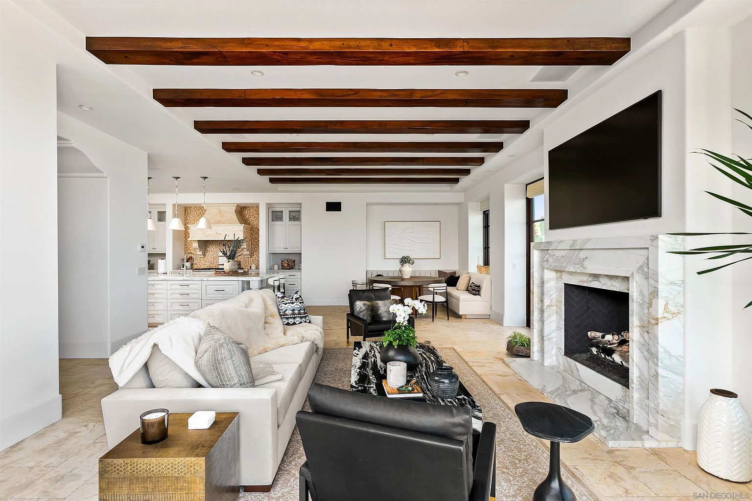 This open-concept living space features a prominent stone-clad fireplace surround and a series of dark wood ceiling beams that draw the eye across the room. The area is furnished with a plush light-colored sofa, two black leather armchairs, and a striking black-and-white patterned coffee table. In the background, a kitchen with light cabinetry and a dining nook with a built-in bench create a cohesive, airy living environment.
