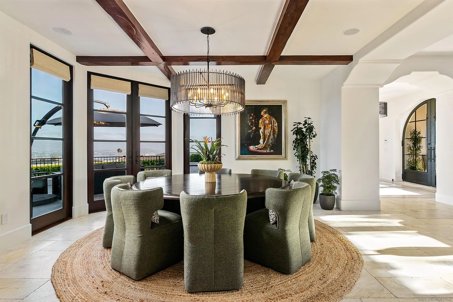 This elegant dining room features a large, round dark wood table surrounded by six upholstered green chairs set atop a circular jute rug. A striking, multi-tiered cylindrical chandelier hangs from a ceiling accented with dark wood beams, while large glass doors provide ample natural light and views of the outdoors. The space is finished with neutral walls, light-toned tile flooring, and a piece of framed artwork, creating a sophisticated and inviting atmosphere.