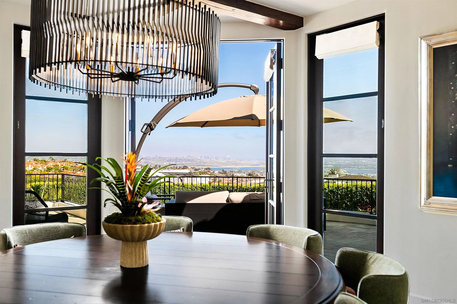This elegant dining area features a large, dark wood circular table centered beneath a striking, modern cylindrical chandelier with vertical slats. The room is framed by tall windows and glass doors that open onto a balcony, showcasing a panoramic view of the city and coastline. The space is decorated with a vibrant floral centerpiece, creating a sophisticated and inviting atmosphere for dining with a view.
