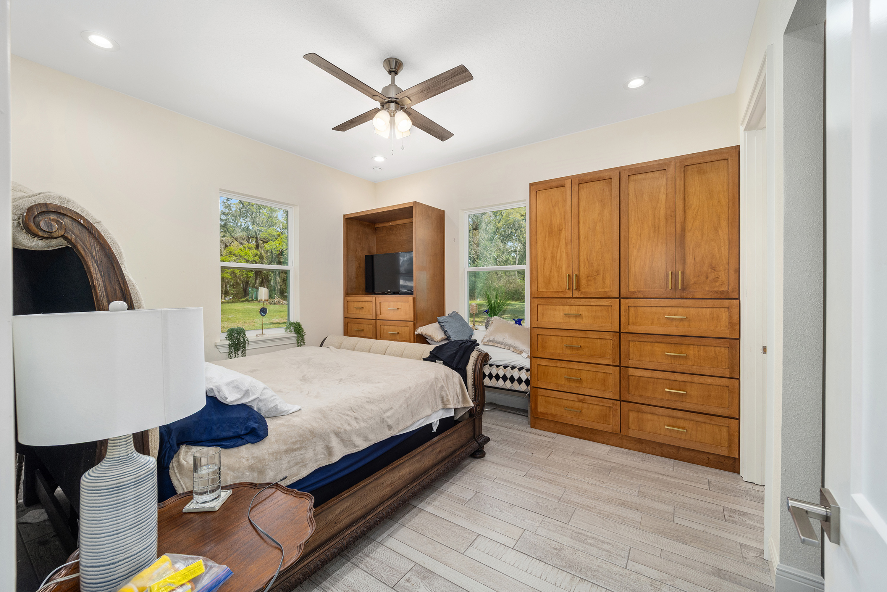 This primary bedroom showcases a blend of traditional and contemporary styles. The room features a large wooden bed, built-in wooden cabinetry, and a window seat nestled between the cabinetry and a window. The light wood flooring and neutral wall color create a bright and inviting atmosphere.