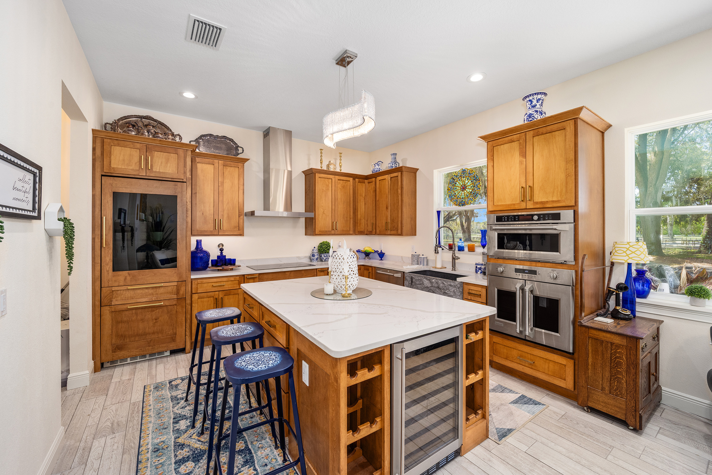 This is a well-appointed kitchen featuring warm wood cabinetry, stainless steel appliances, and a spacious island with a white countertop. The island also includes a wine cooler. The kitchen benefits from natural light through a window overlooking the outside, and is styled with blue and white decorative accents.