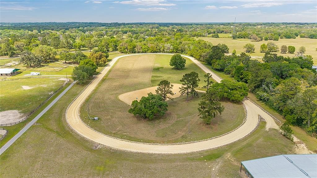 This aerial shot showcases a horse training facility with a well-maintained oval track surrounded by lush greenery and mature trees. The track's sandy surface contrasts with the vibrant green grass and the distant treeline, creating a visually appealing scene. A few buildings are visible in the distance, suggesting a functional and private estate.