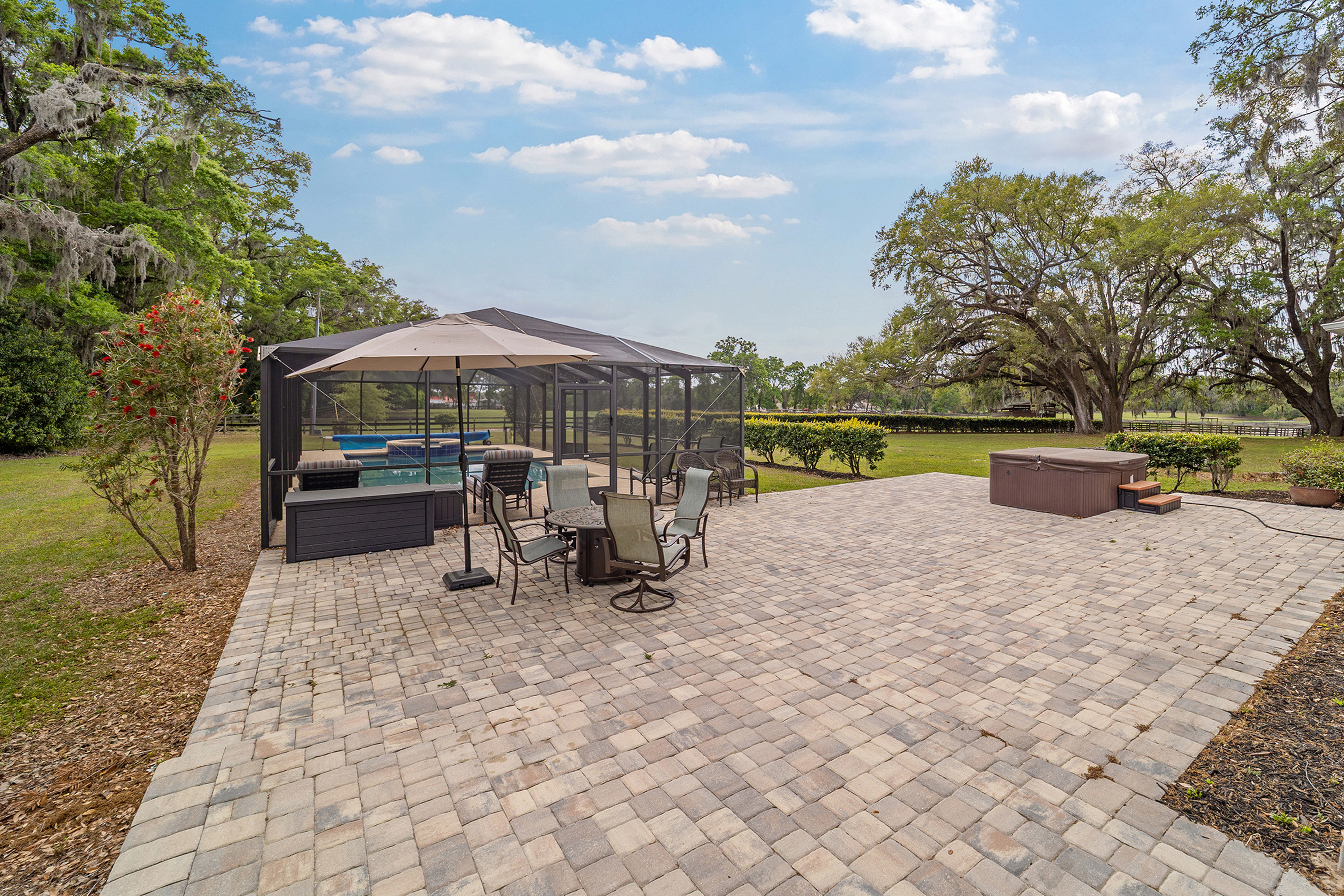 This exterior image showcases a spacious patio area perfect for outdoor living and entertaining. The patio features attractive brick pavers, a covered pool, and a dining set. Adjacent to the pool, a hot tub offers additional relaxation opportunities, all set against a backdrop of lush greenery and mature trees.
