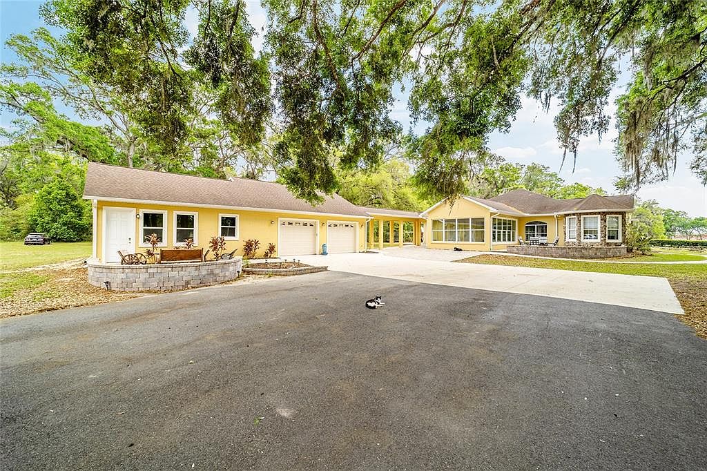 This is a front view of a charming yellow house with a well-maintained asphalt driveway. The house features multiple garage doors, a connecting covered walkway, and a stone-accented facade. Mature trees with Spanish moss provide shade and add to the property's appeal.