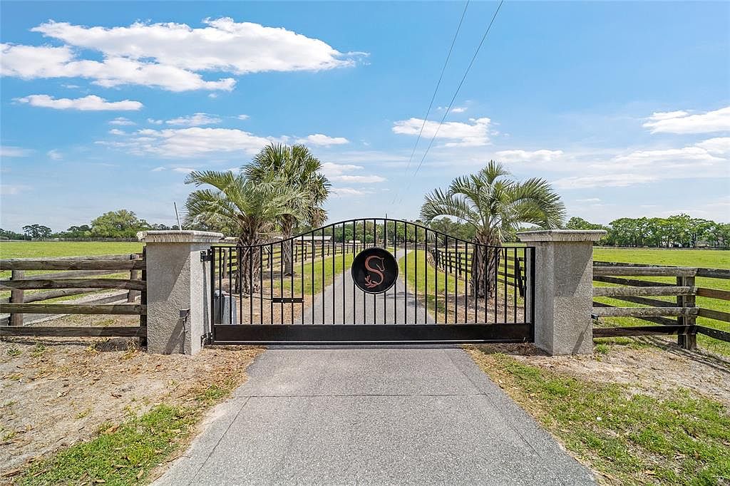 The image showcases a grand entryway to a property, featuring a black metal gate adorned with a circular emblem. The gate is flanked by substantial concrete pillars and wooden fencing, leading to a paved driveway. Palm trees are visible behind the gate, adding a touch of tropical elegance to the estate.