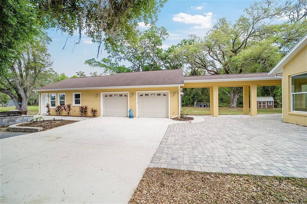 This is a front exterior view of a single-story home with a two-car garage. The house is painted in a warm yellow tone, complemented by a brown roof and light-colored garage doors. A covered walkway connects the garage to the main house, and the driveway is a combination of concrete and brick pavers. Landscaping includes small shrubs and trees, adding to the property's curb appeal.