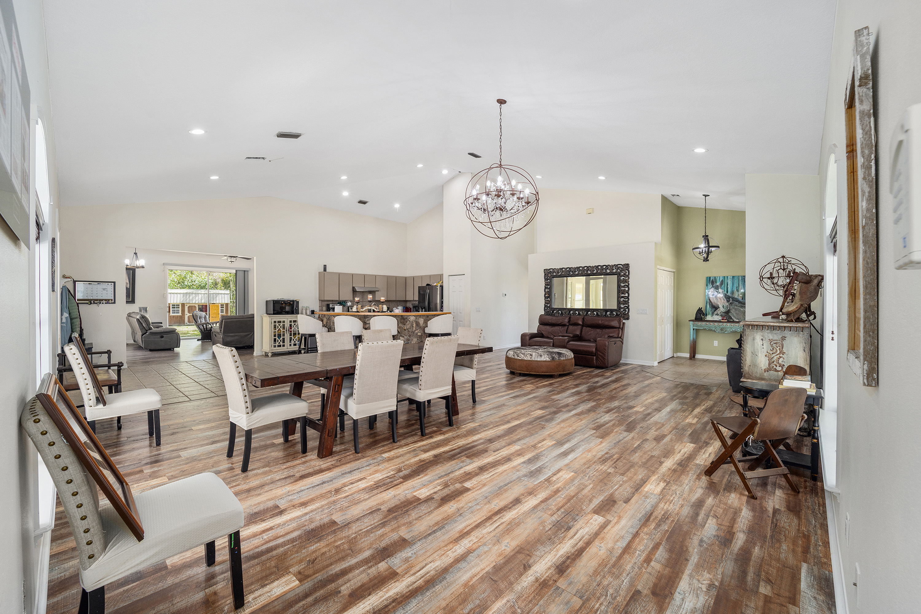 This interior shot showcases a spacious dining area with a rustic-chic aesthetic. A large wooden dining table is surrounded by upholstered chairs, and a unique chandelier hangs above. The room features wood-look flooring and connects seamlessly to the adjacent living areas, creating an open and inviting space for entertaining.