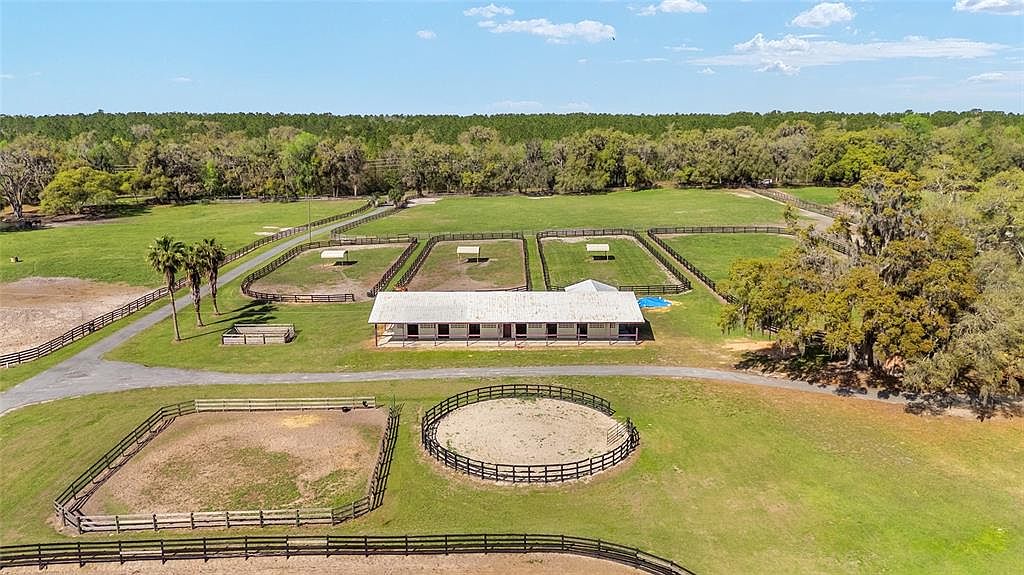 This aerial view showcases a sprawling horse farm with multiple fenced paddocks, a central stable building, and a riding arena. The property is surrounded by lush green fields and mature trees, creating a serene and private setting. A long driveway leads to the stable, emphasizing the scale and grandeur of the estate.