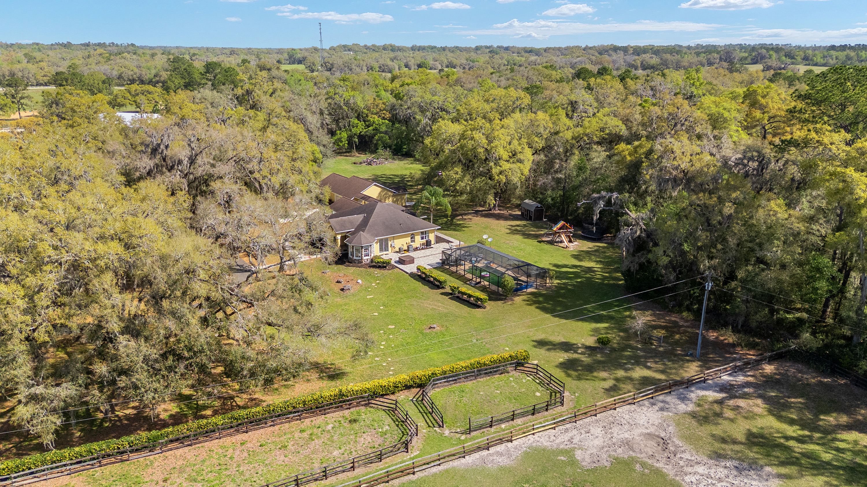 This aerial shot showcases an expansive property featuring a yellow house with a dark roof, surrounded by lush greenery and mature trees. The backyard includes a screened-in pool, a playground, and fenced enclosures, suggesting ample space for recreation and outdoor activities. The property's size and landscaping offer a sense of privacy and a country estate feel.