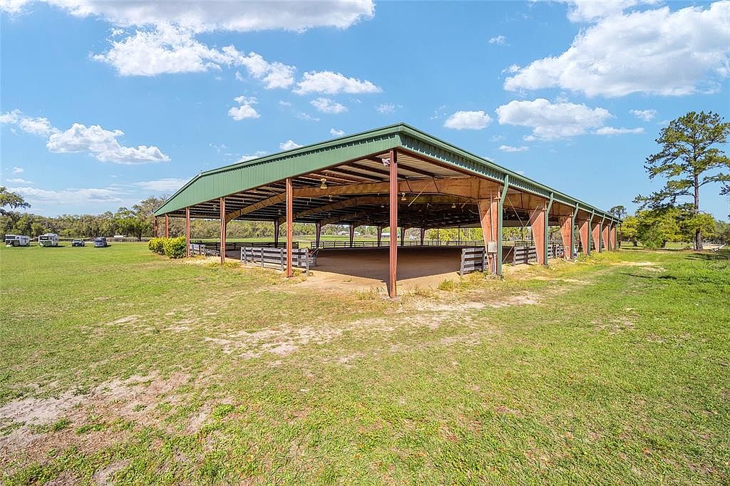 The image showcases a large, well-maintained outdoor riding arena with a green metal roof supported by sturdy posts. The arena is surrounded by a wooden fence and lush green grass, creating a spacious and inviting environment. In the background, trees and a few vehicles can be seen, adding depth to the scene.