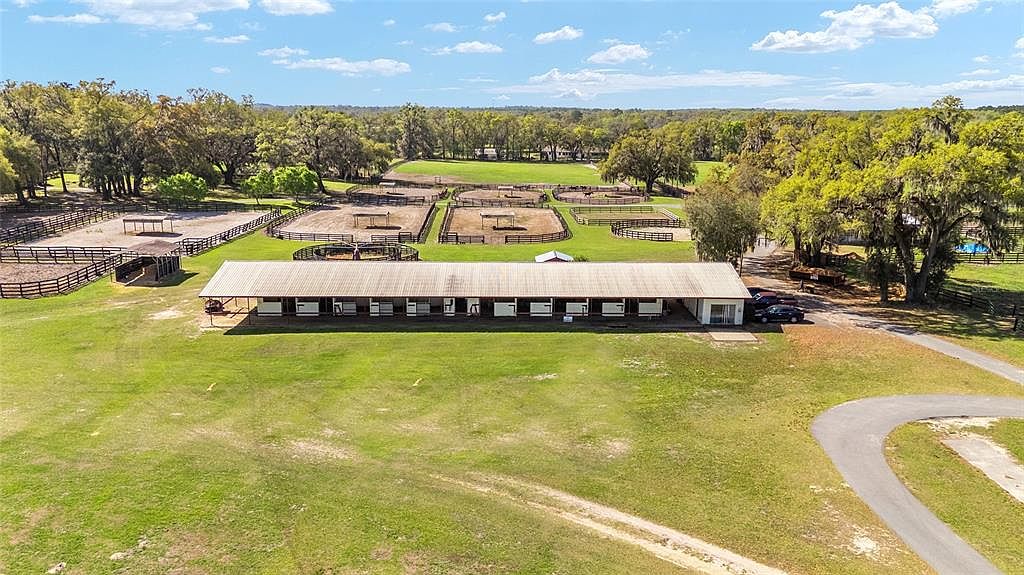 This aerial shot showcases a sprawling equestrian estate. The property features multiple fenced paddocks, a long stable building with numerous stalls, and a well-maintained green lawn. A winding driveway leads to the stable, and mature trees provide shade and privacy, creating a picturesque and functional horse farm.