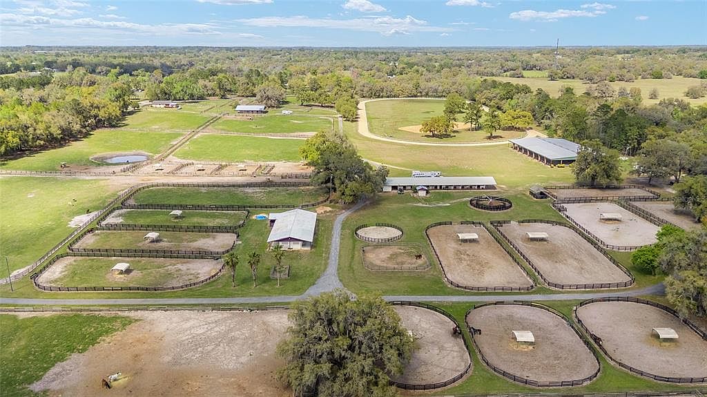 This aerial shot showcases a sprawling horse farm with multiple paddocks, barns, and riding arenas. The property features lush green pastures, mature trees, and well-maintained facilities, creating an idyllic setting for equestrian activities. The perspective is high-angle, providing a comprehensive view of the entire estate.