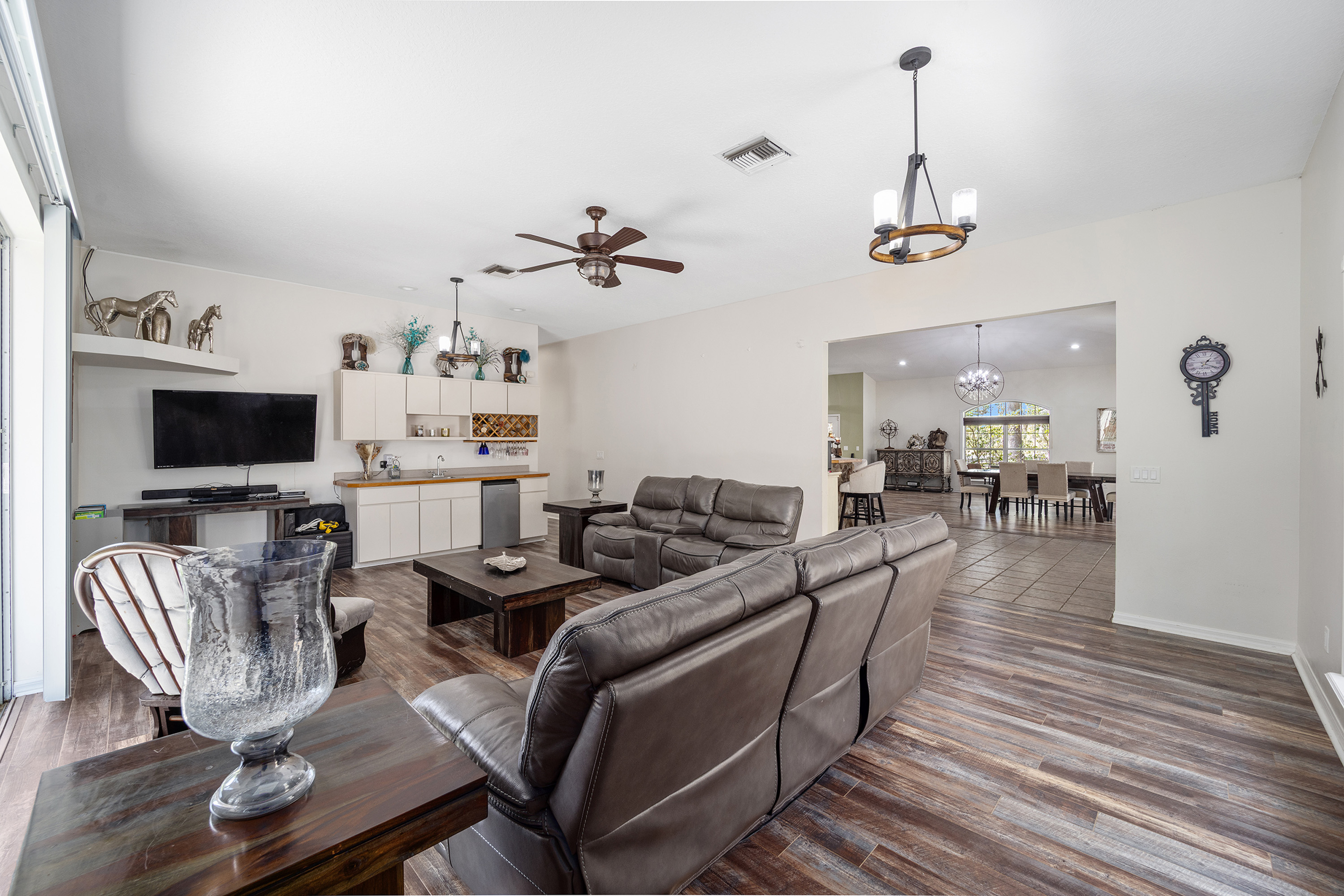 This is a well-lit living room featuring two leather reclining sofas, a dark wood coffee table, and a custom-built wet bar. The flooring is wood-look laminate, and the room opens into a dining area, creating an open-concept living space. A ceiling fan and decorative light fixtures add to the ambiance of the room.